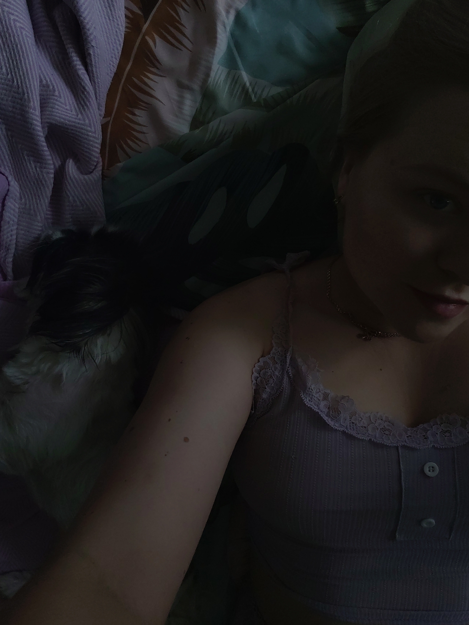 A woman is lying on a bed with a dog. She is wearing a lilac tank top with lace trim and a necklace. The bed has several different patterned blankets. The photo appears to be taken indoors in a dimly lit room. There is no indication in the image that it was taken in Izhevsk, Russia.