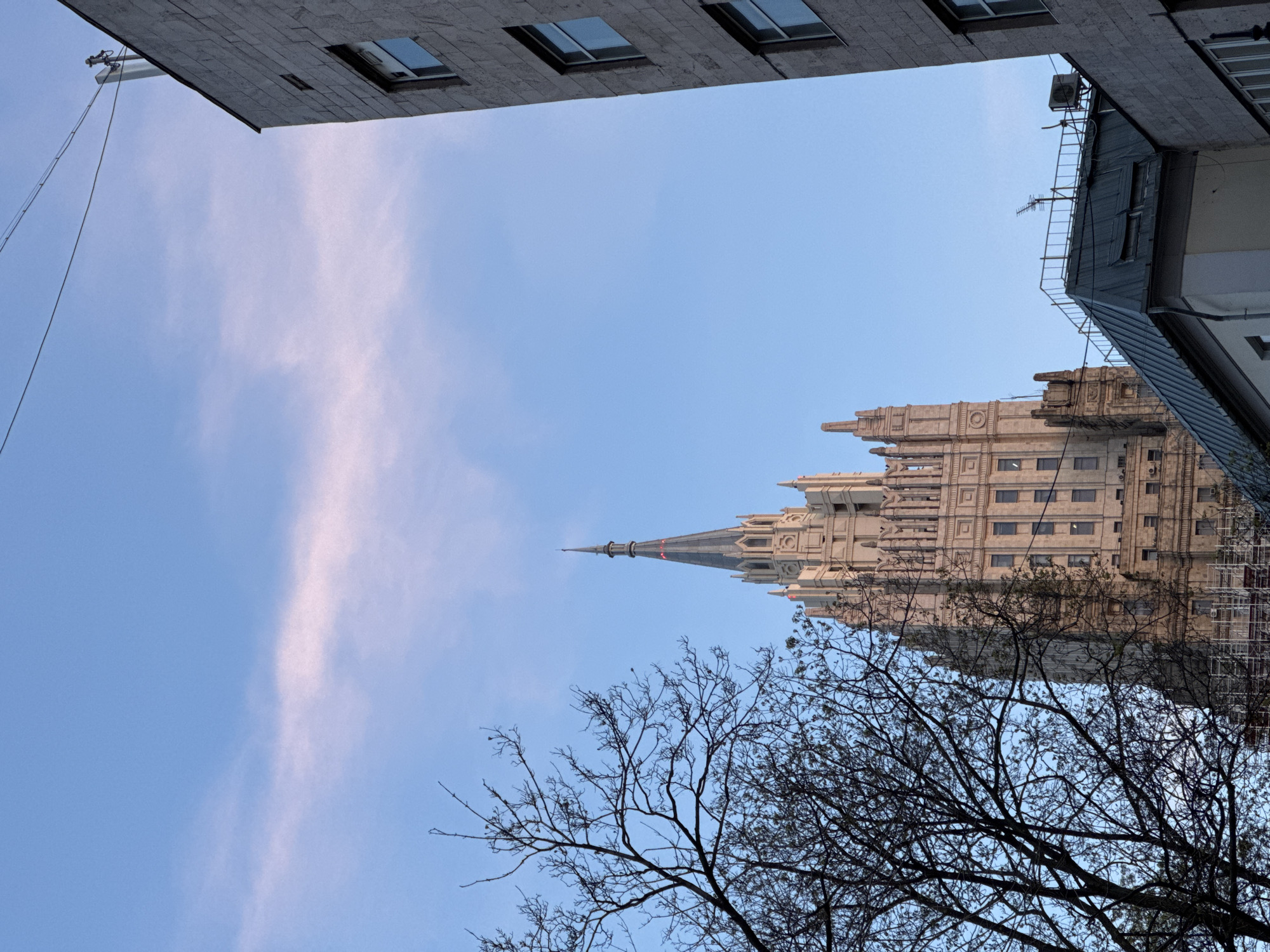 The image shows a view upwards in Moscow, Russia, showcasing the Ministry of Foreign Affairs building. The perspective is from street level, looking up between two buildings; one is modern with rectangular windows, and the other is older with a sloped roof and architectural details. The Ministry building is a large, ornate structure with a spire. Bare tree branches are in the foreground against a blue sky with light clouds. Scaffolding is visible on the side of the building, indicating ongoing maintenance or construction. There are no people visible in the frame.