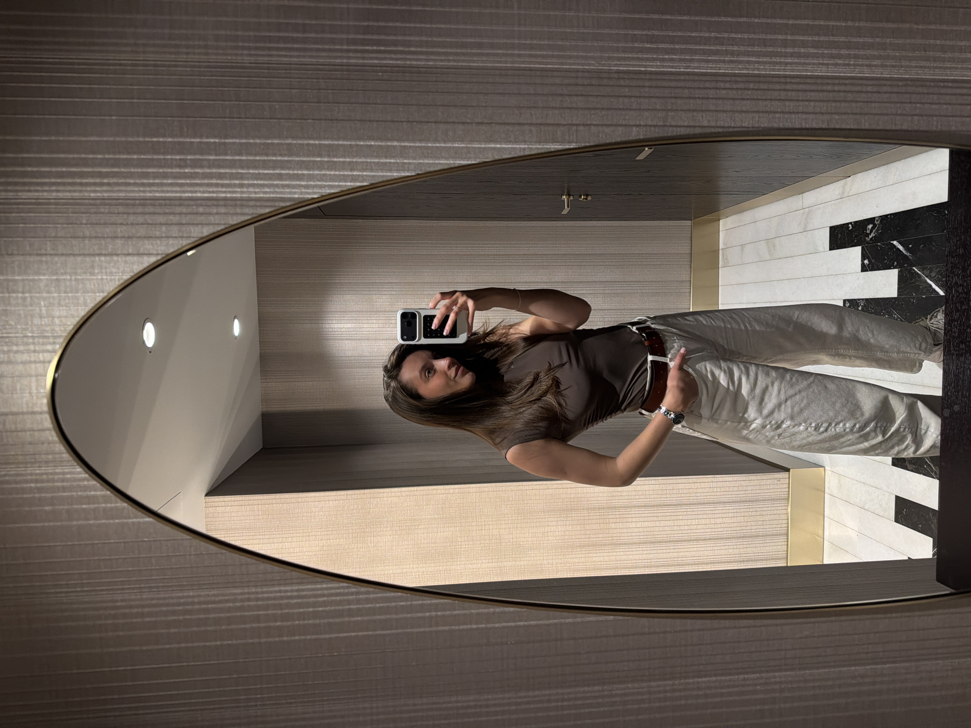 A full-body selfie of a woman is reflected in an oval mirror. The woman has long brown hair and wears a brown top and light-colored pants with a brown belt. She holds a white phone with a black lens protector. The mirror is framed by a gold trim and is mounted on a horizontally striped wall. The reflection reveals a tiled wall and floor, including black and white geometric patterns. The scene is likely in a bathroom or fitting room. The location is unknown.