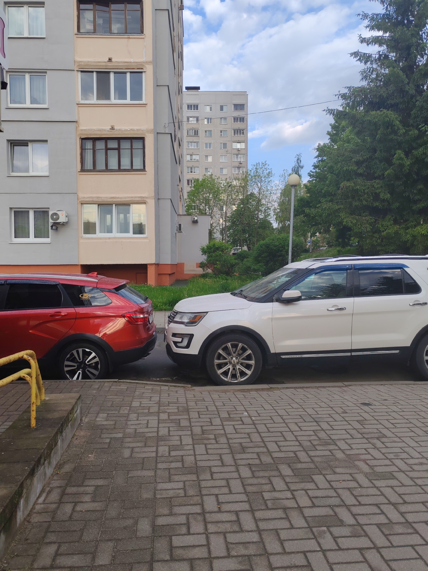 The image shows two cars parked very close to each other on a brick-paved area alongside a building. The car on the left is red, while the one on the right is white, and the cars look as if they are touching. A yellow guardrail is visible near the red car. The buildings are multi-story with repeating window patterns, suggesting an urban residential area. The sky is partly cloudy. The presence of trees and streetlights suggests a developed area, potentially a city. The location is the city of Unknown, Unknown.