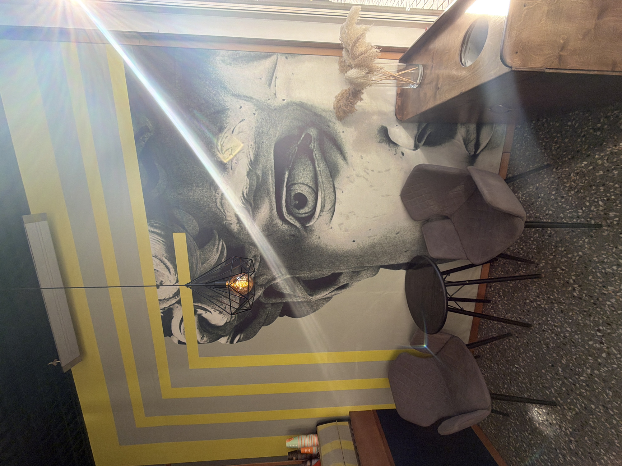 The image depicts the interior of a room. The room features a wall decorated with a large black and white image of a classical sculpture, possibly a bust or head. The wall is also adorned with horizontal stripes in shades of gray and yellow. A modern, geometric-style pendant light hangs from the ceiling. Two modern-style chairs with gray upholstered seats are positioned next to a small round table with a dark surface. The floor appears to be a terrazzo or speckled material. On a shelf next to the striped wall are a stack of orange and white patterned cups or containers. A wooden desk or counter surface is partially visible at the top of the frame, with a small vase holding dried plants. The image is shot from a slightly elevated angle and is rotated, making it appear as if the room is sideways.
Based on the decor and architectural style, it is likely the location is a cafe or restaurant in a modern urban setting. It is impossible to confirm the location being in Krasnoyarsk, Russia from the visual data alone.