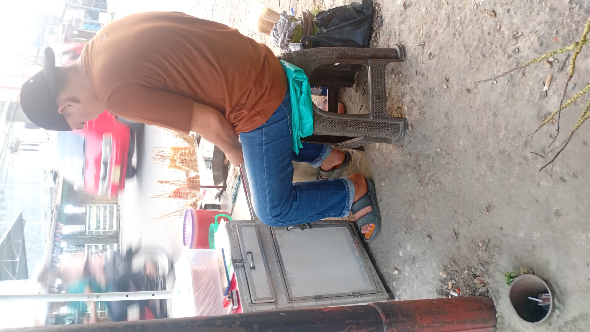 The image depicts a street vendor in Jambi, Indonesia, preparing food. A man wearing a brown t-shirt, blue jeans, and black sandals is seated on a brown plastic stool with a teal plastic bag on top, facing away from the camera. He is positioned next to a silver food cart with a white panel and a drawer. Behind him, on the cart, are various food items on skewers, plastic containers, and a smartphone. The street appears to be made of concrete and dirt, with scattered debris. A red vehicle is blurred in motion in the background, suggesting a busy street. To the left of the cart, a red pole stands vertically. In the foreground, a circular drain opening contains a drink carton. The overall scene suggests a typical roadside food stall environment in an urban setting.