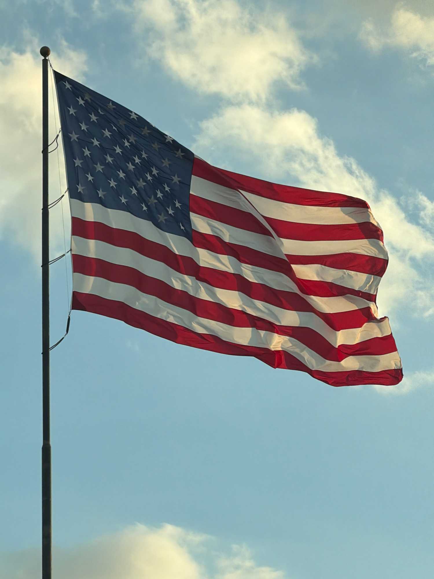 An American flag is shown unfurled and waving in the wind, attached to a tall, dark flagpole. The flag is positioned on the left side of the frame, taking up a significant portion of the image. The sky behind the flag is a pale blue, with scattered white clouds that appear bright, suggesting either daylight or the sun is positioned behind them. The flag's stripes and stars are clearly visible. The flagpole is a dark, cylindrical object with a spherical finial at its top. There are no people or other objects visible in the image.