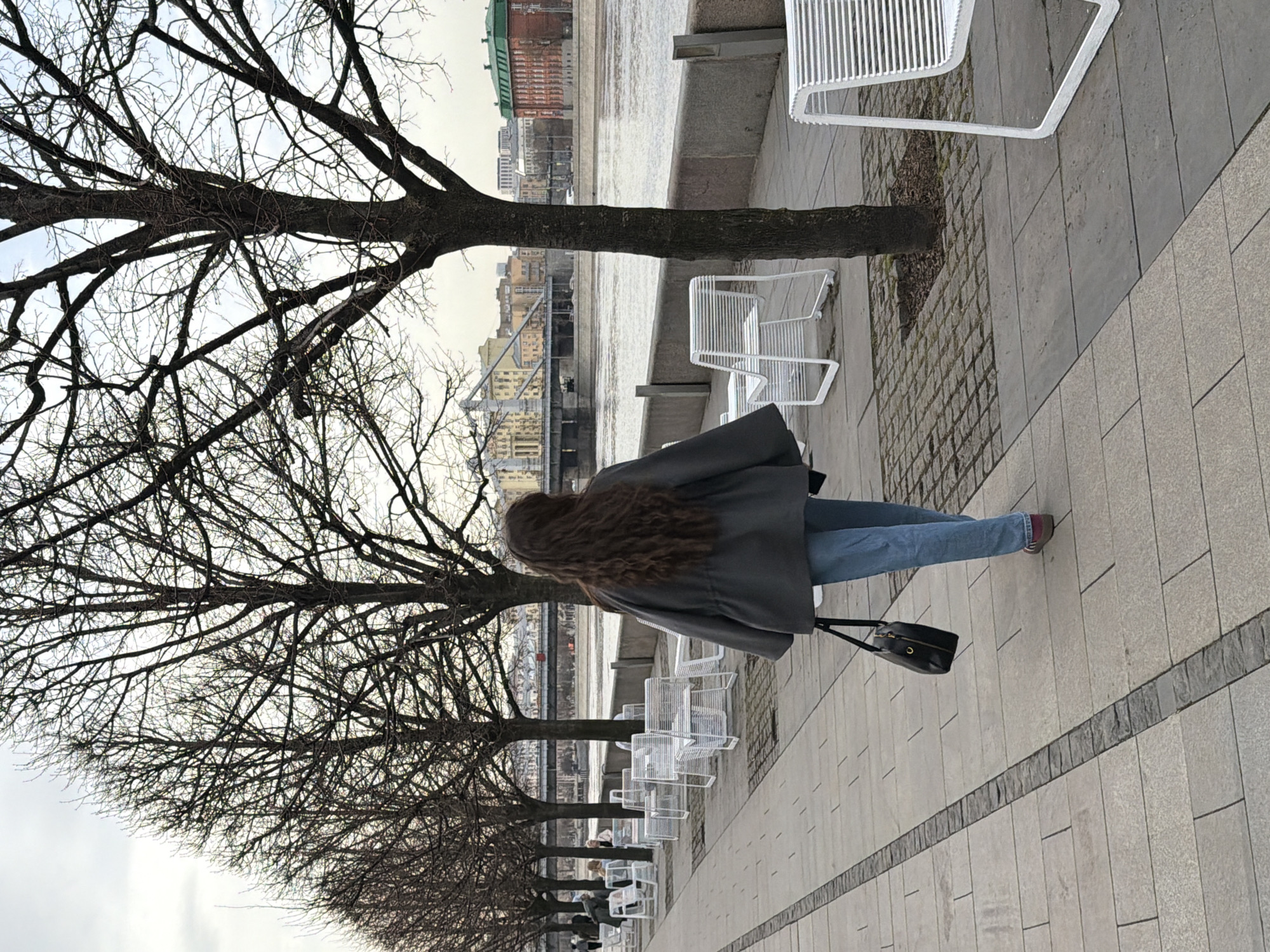 The image shows a woman walking along a paved walkway in Moscow, Russia. She has long brown hair and is wearing a gray jacket and blue jeans. She is carrying a black handbag. The walkway is lined with bare trees and white metal chairs. In the background, there is a river and buildings. The sky is overcast.