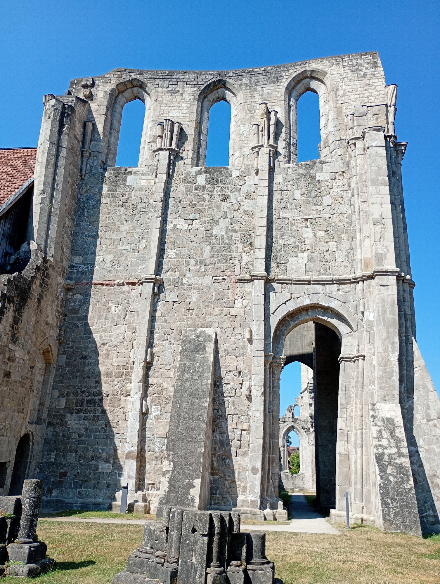 This is a low-angle, full-frame, eye-level shot of the ruins of the former Walkenried Abbey in Walkenried, Germany. The dominant subject is the large, weathered stone facade of the abbey, characterized by its rough-hewn stone blocks and crumbling architectural features.
The upper section of the facade features a series of arched openings, most of which are empty, revealing a clear blue sky. The stonework is varied, with some blocks showing signs of erosion and discoloration. Vertical, pilaster-like elements are integrated into the wall.
Below the upper arches, a large, intact arched doorway is prominent, leading into a darker interior. Through this doorway, another, smaller arched opening is visible in the distance, further within the ruins. To the left of the main facade, a section of a red-tiled roof is visible, suggesting the presence of a partially preserved or reconstructed building adjacent to the ruins.
The ground in front of the ruins is covered in dry, yellowed grass. In the foreground, several weathered stone fragments, resembling broken columns or decorative elements, lie scattered on the ground. The lighting indicates a bright, sunny day, with strong shadows cast by the architectural features. The sky is a vivid, cloudless blue. There are no people visible in the image.