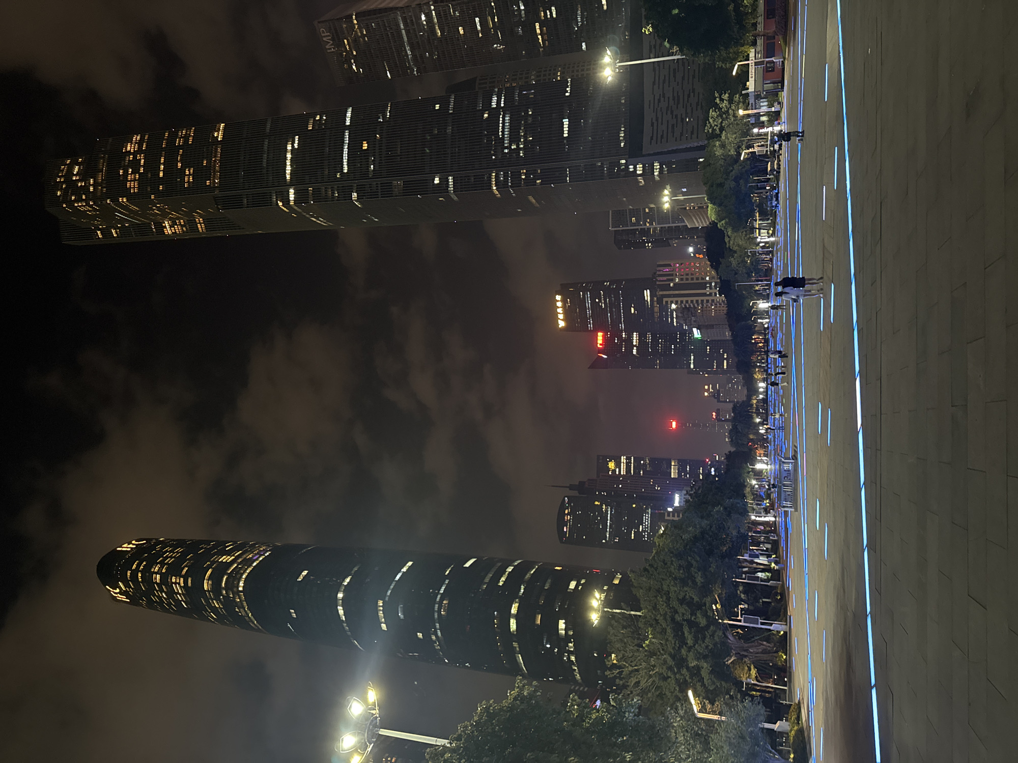 The image depicts a nighttime cityscape in the Tianhe District of Guangzhou, China. Skyscrapers dominate the skyline, illuminated with numerous lights. Below, a pedestrian walkway paved with light blue features spans the scene. Several individuals are present, walking or standing on the walkway. Vegetation, including trees, is interspersed between the buildings and the walkway. The overall environment is urban, with a modern architectural aesthetic.