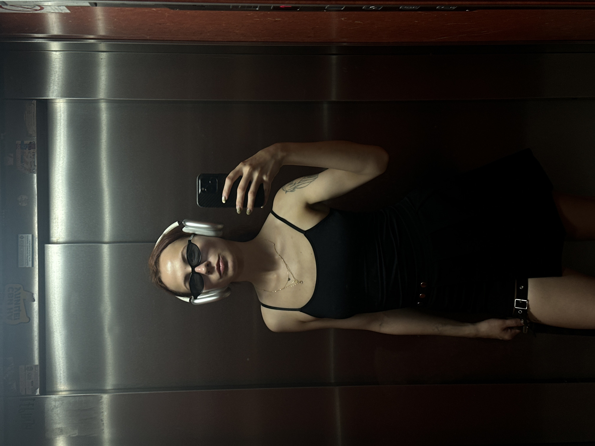 A young woman is taking a selfie in what appears to be an elevator. She is wearing a black dress, sunglasses, and white headphones. She is holding a black smartphone in her right hand. The elevator has a metallic surface and appears to be located in Novi Sad, Serbia, based on signage partially visible on the left side of the image.