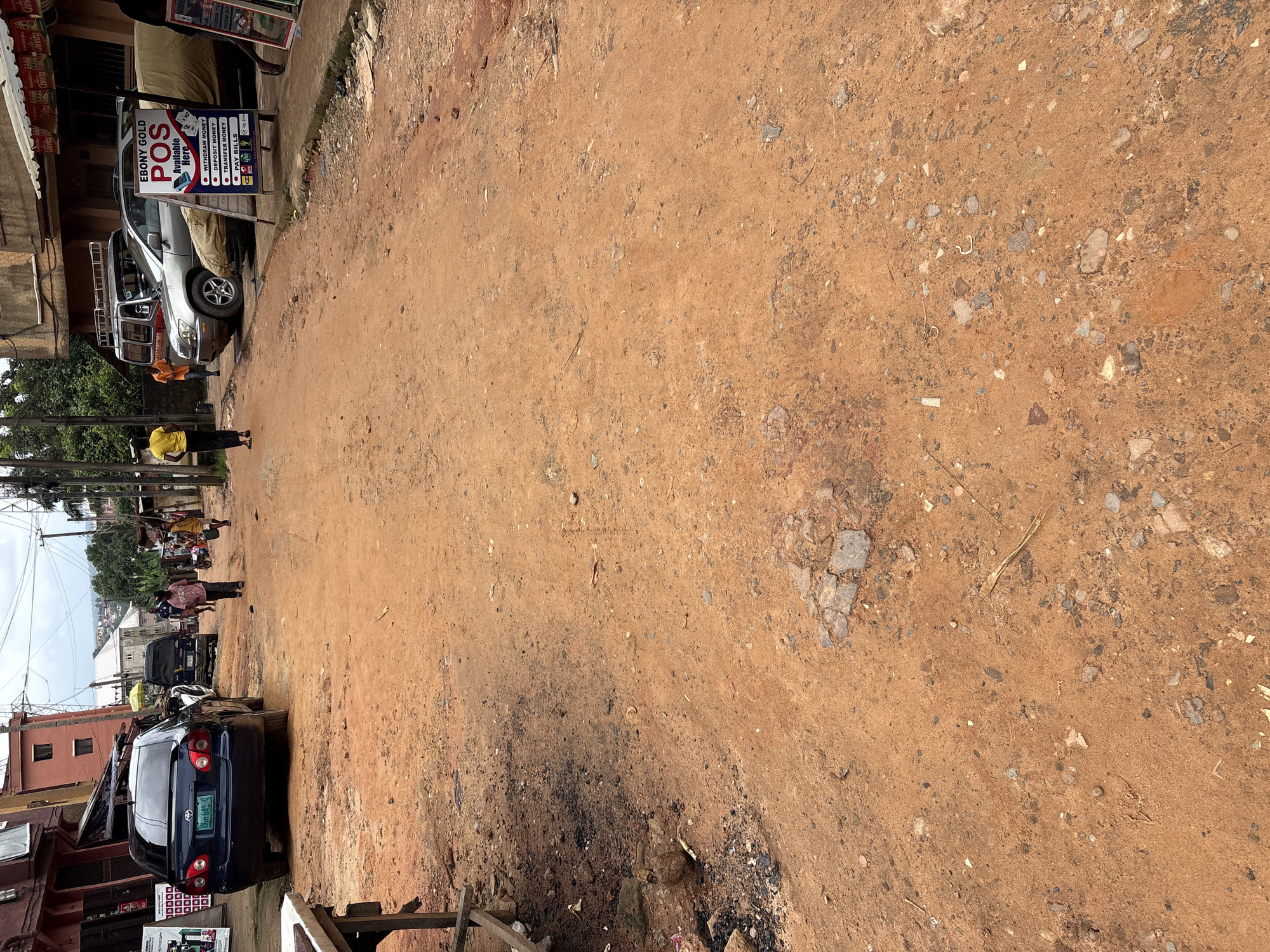 The image depicts a street scene in Nkpor, Nigeria, characterized by a dirt road with patches of gravel and debris. On the left side of the road, a dark blue sedan is parked, displaying a license plate that reads "UGH 649 JK". Further down the road, a silver SUV is visible. A few individuals are present: one person wearing a yellow shirt walks away from the camera, and another in an orange shirt is walking towards a parked van. A poster advertising "EBONY GOLD POS" services, listing options like "WITHDRAW MONEY", "DEPOSIT MONEY", "TRANSFER MONEY", and "PAY BILLS", stands beside the silver SUV. The surrounding environment consists of buildings, some with multiple stories, and overhead power lines. The overall impression is that of a busy, somewhat unpaved urban thoroughfare.