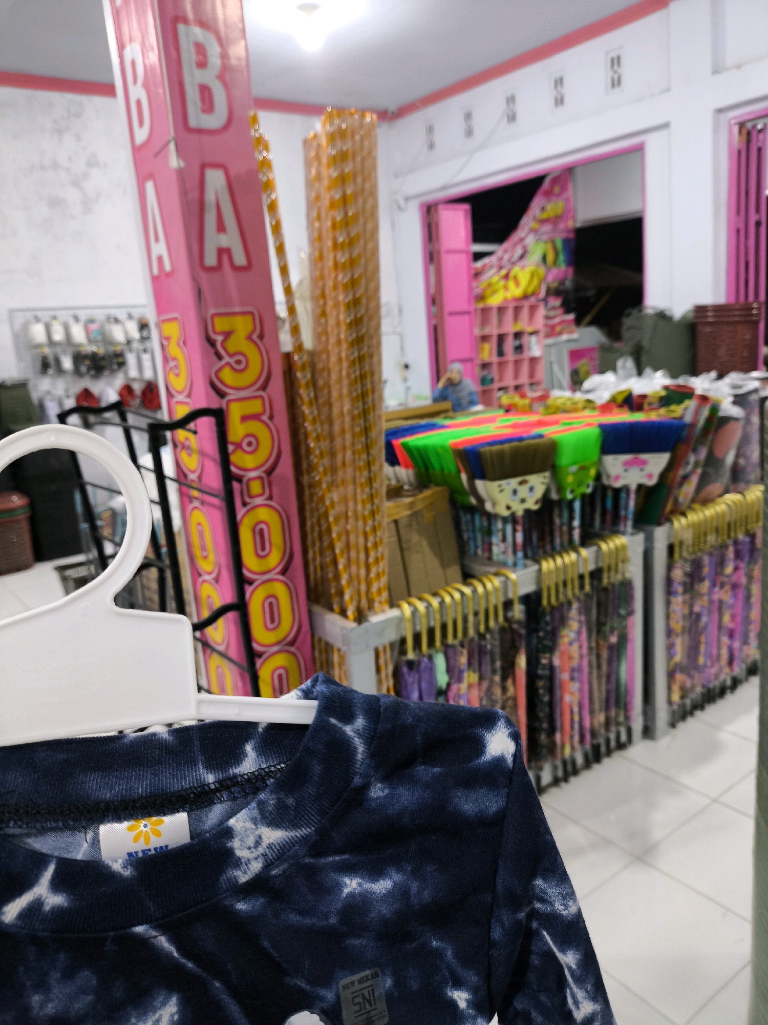 The image depicts the interior of a shop in the city of Posso, Indonesia, featuring various items for sale. In the foreground, a dark blue tie-dye t-shirt is displayed on a white hanger. Behind the t-shirt, a tall pink banner with white lettering "BBA" and yellow numbers "335.000" is visible. To the right of the banner, there are stacks of yellow and orange striped items, possibly straws or decorative elements. Further into the shop, rows of colorful brooms with green, blue, and red bristles are arranged on a display. Below the brooms, numerous items, likely clothing or fabric, are hanging on racks. In the background, a person wearing a blue hijab is partially visible near a pink shelf unit filled with items. The shop appears to have a tiled floor and fluorescent lighting overhead.