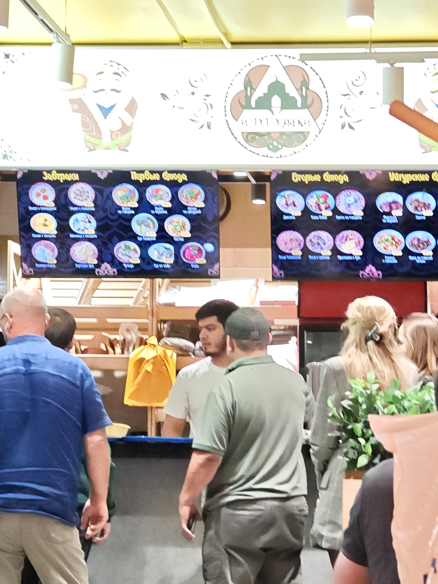 The image shows a food court setting with a menu board displaying various dishes, categorized into "Завтраки" (Breakfasts), "Первые блюда" (First courses), and "Вторые блюда" (Second courses), and "Узбекские блюда" (Uzbek dishes). The menu board features pictures of food items with their names below. Several people are visible in the foreground, including a man in a blue shirt facing away from the camera, another man in a green shirt facing sideways, and a woman with blonde hair. Behind them, there is a food stall with wooden shelving and a yellow bag. The overall environment suggests a casual dining area.