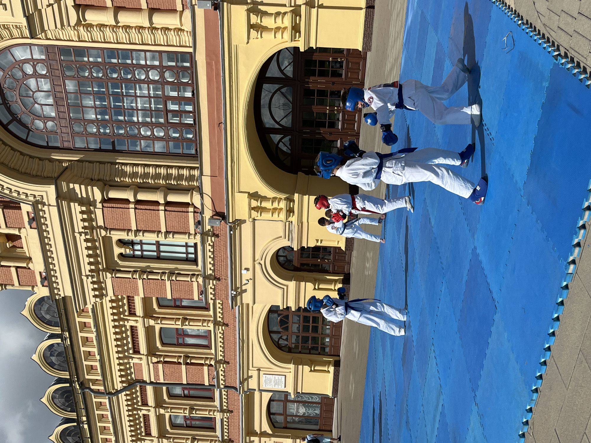 In Nizhny Novgorod, Russia, on a sunny day, several children in white karate gis with blue and red belts and protective headgear are practicing martial arts on a blue tatami mat in front of a grand, yellow and red brick building with arched doorways and large windows. The building's facade features intricate architectural details, including decorative moldings and columns. Some children are sparring, with one child in a blue headguard throwing a punch towards another, while others observe or prepare for their turn. The scene is set outdoors, with the pavement visible around the tatami mat.