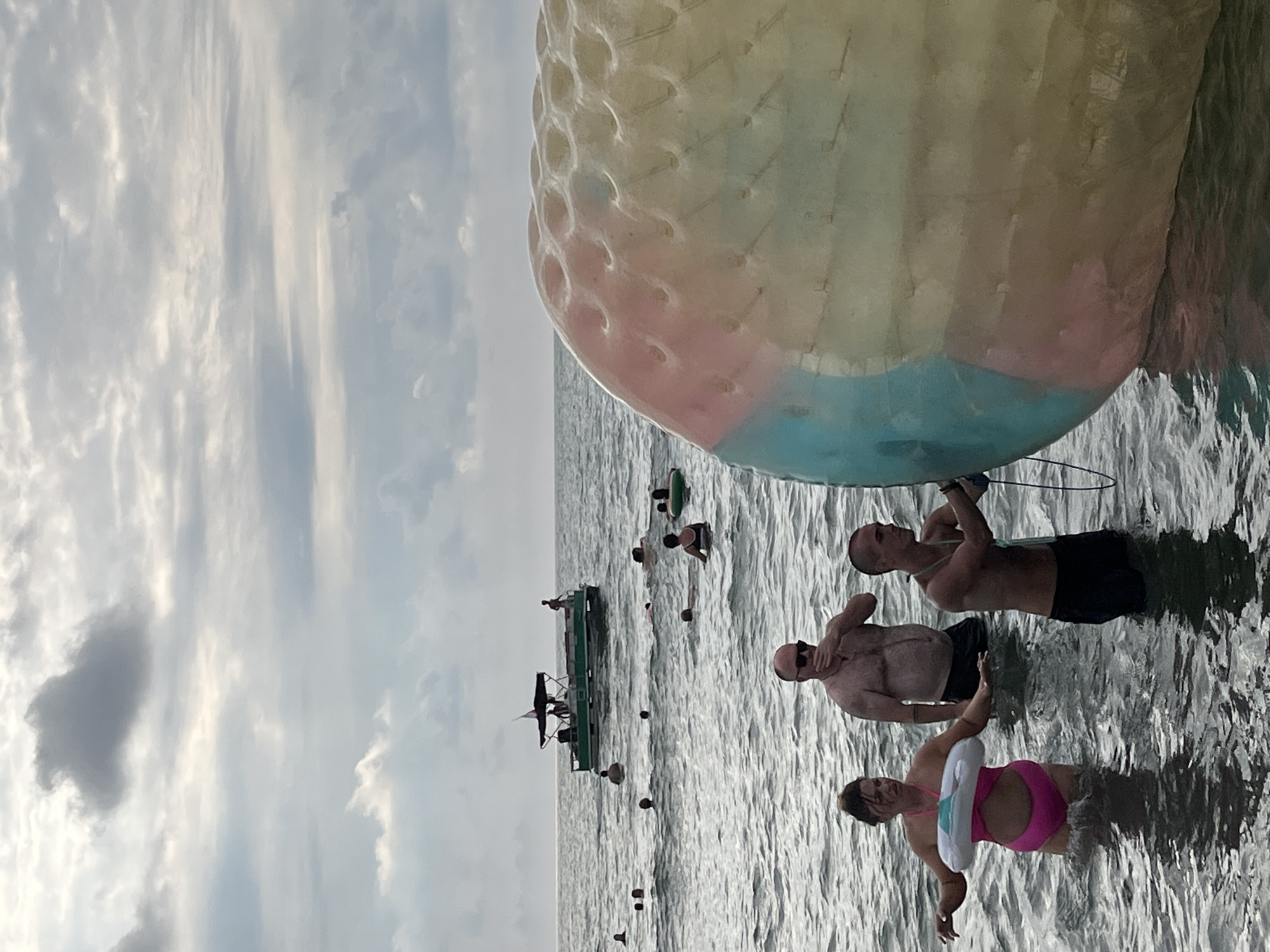 In Ureki, Georgia, on a cloudy day, people are wading in the Black Sea. A large, inflatable, multi-colored sphere, likely a zorbing ball, dominates the foreground on the right. In the mid-ground, three individuals stand in the water. A woman in a pink swimsuit with a white inflatable ring around her waist is on the left, facing forward with arms extended. To her right, a man in sunglasses and swim trunks is touching his face. Further to the right, a man in dark swim trunks is holding a rope attached to the inflatable sphere and looking to his right. In the background, a green pontoon boat with a canopy is visible, carrying several passengers. Other people are scattered throughout the water, swimming and relaxing. The sea is calm with visible ripples. The sky is overcast with layers of grey and white clouds.