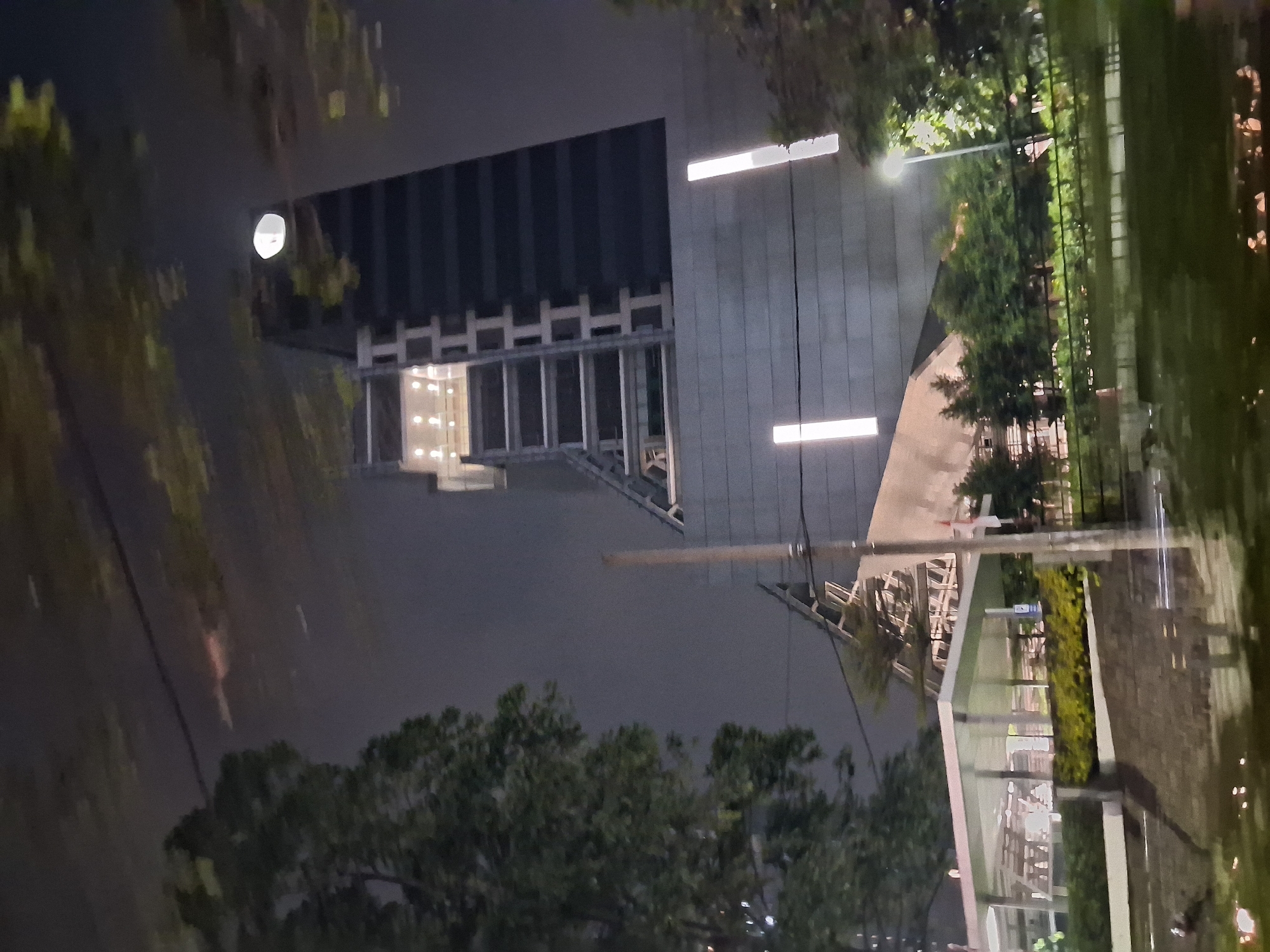 The image, taken at night in Depok, Indonesia, shows a modern building with a dark facade and a lighter-colored section under construction. The construction area has exposed beams and scaffolding. In front of the main building, there is a covered structure with a white roof, supported by pillars, and surrounded by green landscaping. The scene is illuminated by artificial lights, including vertical strip lights on the building and streetlights. Trees are visible in the foreground and around the structures. A faint reflection on a wet surface, possibly a road or a pond, is visible at the bottom of the image. A bright, round light, possibly the moon or a distant lamp, is visible in the sky. The overall impression is a contemporary urban environment at night, with a mix of completed and under-construction structures.
