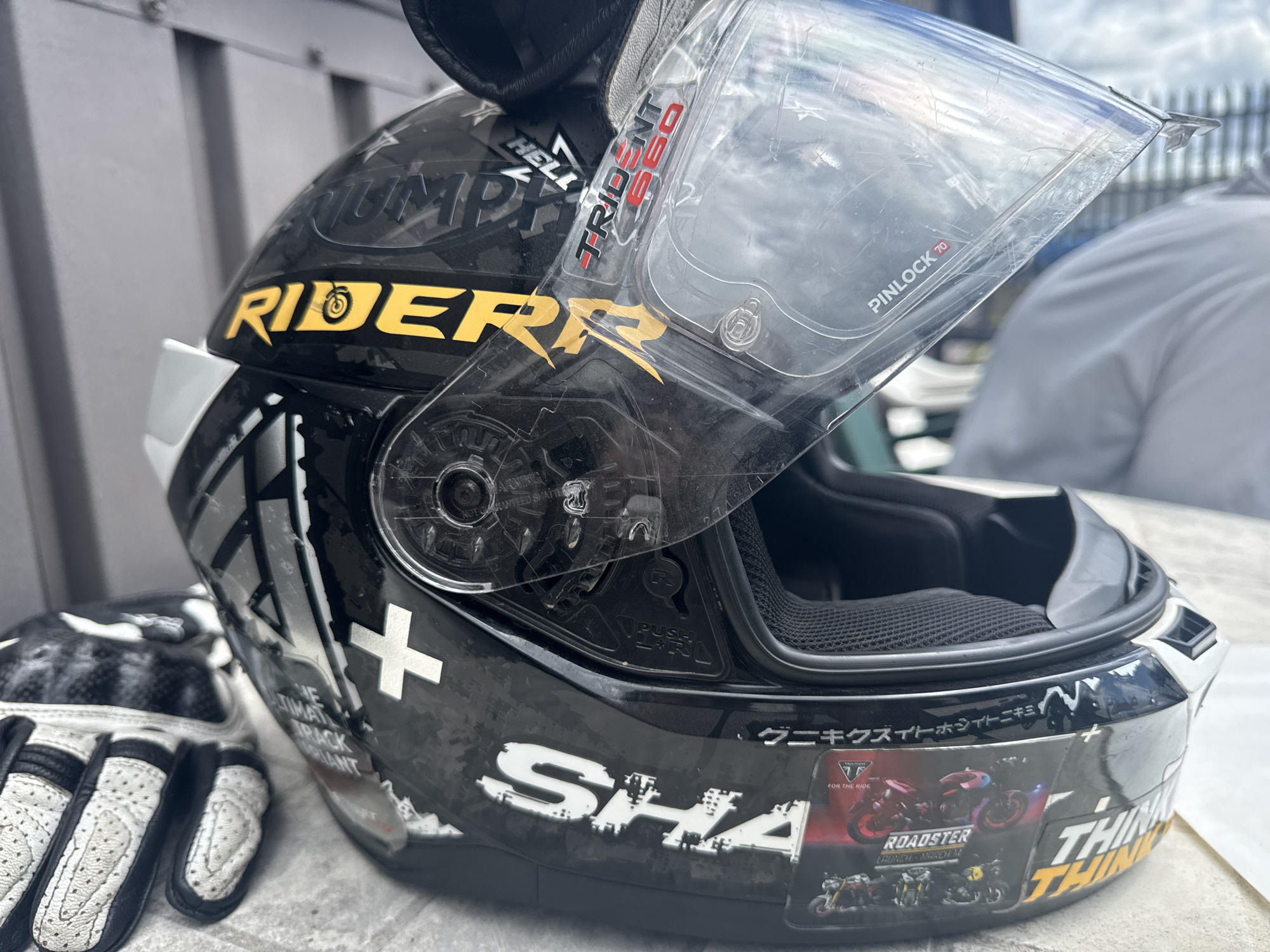 A black motorcycle helmet with a clear visor is resting on a table outdoors. The helmet has various decals, including the word "RIDER" in yellow and a "SHARK" logo. A pair of white and black motorcycle gloves is positioned to the left of the helmet. In the background, a person in a grey shirt is visible. The setting appears to be a table outside, possibly in a cafe or seating area in Tewkesbury, United Kingdom, with a metal railing and fence in the distance. The lighting suggests it is daytime, with the sky partially visible through the helmet's visor.