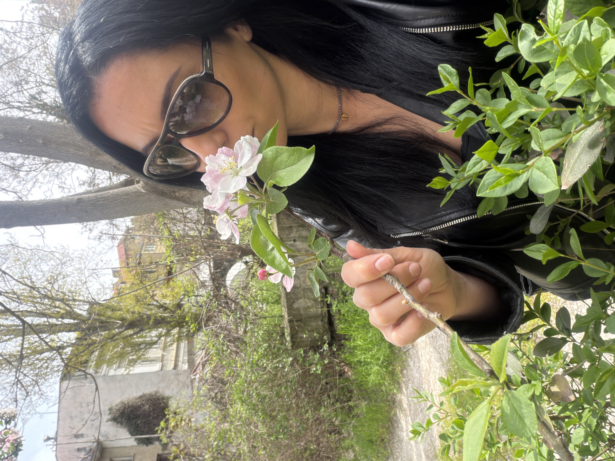 A woman with long, dark hair and sunglasses is seen outdoors in what appears to be a park or garden setting. She is wearing a black leather jacket and holding a branch of flowering blossoms close to her face, as if smelling them. The vegetation around her includes green bushes and trees. A building is visible in the background. Given the metadata of the image, this location could be within the city of Pleven, Bulgaria.