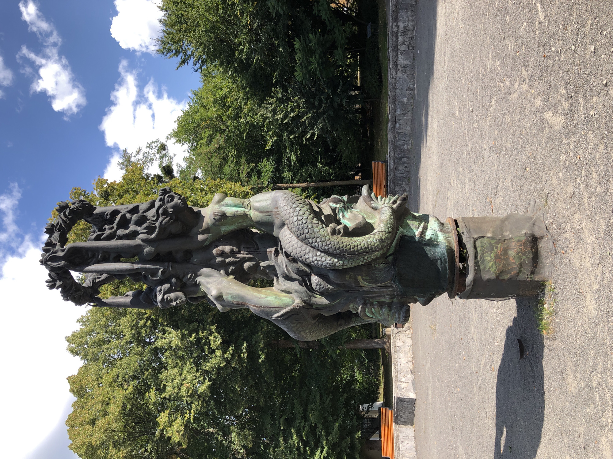 The image shows a bronze sculpture in a park setting. The sculpture is ornate and depicts several figures, including what appear to be mermaids or sirens with fish-like tails, intertwined with a dragon or serpent. The sculpture is weathered and has a greenish patina. It is mounted on a cylindrical pedestal which is partially covered by a dark mesh material. The ground around the sculpture is covered with fine gravel. In the background, there are mature trees with lush green foliage, indicating a summer or late spring season. There are also some park benches visible in the mid-ground, and a stone wall runs along the edge of the gravel area. The sky above is blue with scattered white clouds.
This sculpture is located in Lviv, Ukraine, specifically in the Bohdan Khmelnytsky Park. It is part of a fountain called the "Nymphs Fountain" or "Water Sprite Fountain", a well-known landmark within the park.