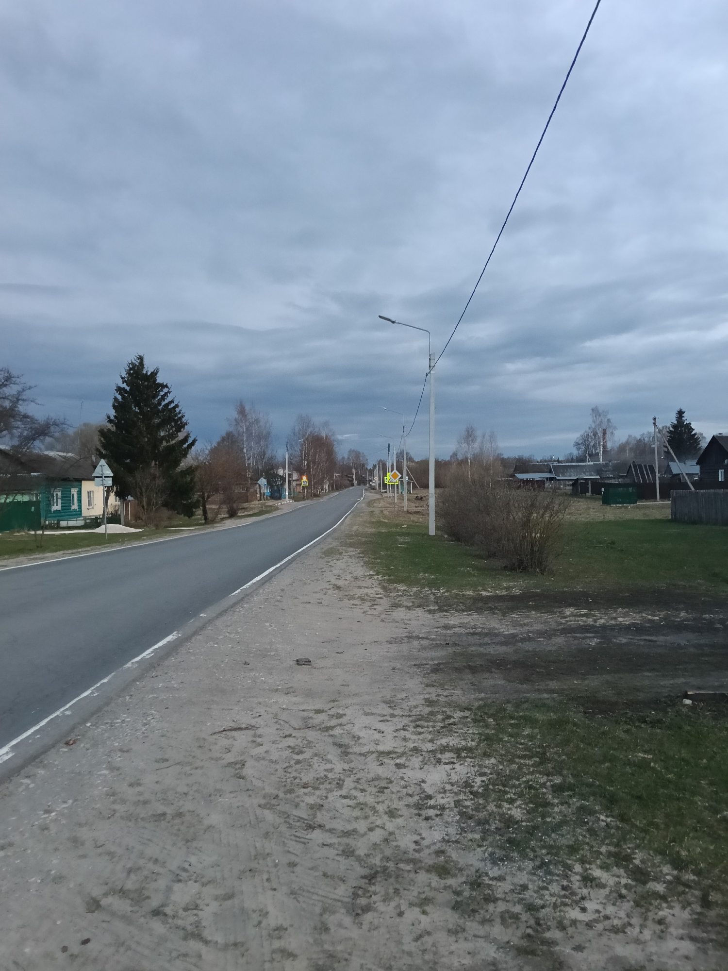 The image captures a quiet, semi-rural road scene in Красный Маяк, Russia, under an overcast, gray sky. The time of day appears to be late afternoon or early evening, or a heavily clouded daylight, resulting in dim and diffused lighting. No people or animals are visible.
The paved road, edged with a solid white line on the right, recedes into the distance, flanked by a dirt and gravel shoulder. On both sides of the road, traditional wooden houses, some painted in green and yellow, are visible, interspersed with trees and bushes. Many trees appear bare or with nascent foliage, suggesting early spring or late autumn.
A prominent utility pole with a streetlight stands on the right side of the road, supporting power lines that crisscross the sky. Further down the road on the right, two yellow road signs are visible: a diamond-shaped sign with a black cross, indicating a crossroads or junction, and a triangular sign with a black walking person, signaling a pedestrian crossing. A small, unreadable white rectangular sign is attached beneath the crossroads sign.
The ground consists of a mixture of dirt, patchy green grass, and some muddy sections. The sky is dominated by uniform gray clouds. The overall scene portrays a still, everyday village environment, characterized by its quietude and rustic charm.