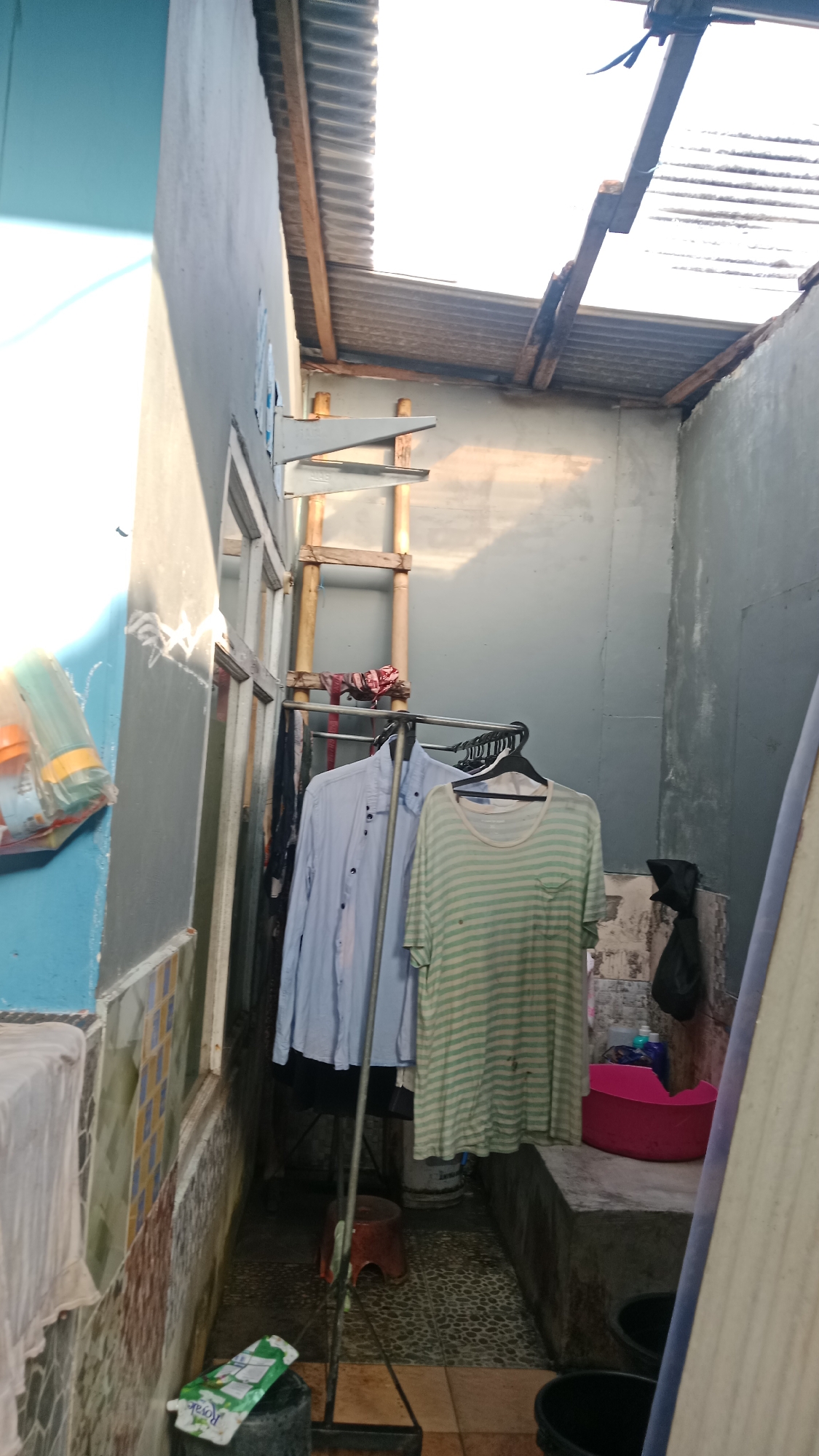 This image depicts an outdoor alleyway in Denpasar, Indonesia, likely a service area or backyard. The scene is characterized by clutter and practical arrangements for daily living. A clothesline, fashioned from a metal rack, is prominently featured, holding several garments including a light blue button-down shirt and a green and white striped t-shirt. Various other clothes are hung on hangers, suggesting recent laundry activity.
The architecture of the area is evident: a textured, light-colored wall forms the backdrop, with a corrugated metal roof supported by wooden beams overhead, indicating a basic outdoor structure. Natural light filters through gaps in the roof, casting shadows. To the left, a blue-painted wall with some tile accents suggests it's part of a dwelling or building.
Objects scattered around the alley include plastic containers, one of which is a bright pink basin. A small red stool is placed near the clothesline. In the foreground, a black bucket and a plastic bag with a green label are visible on the ground, which is paved with patterned tiles. A large, light-colored surfboard or similar flat object leans against the right wall. The overall impression is one of a functional, lived-in space within a residential or commercial setting in Denpasar.