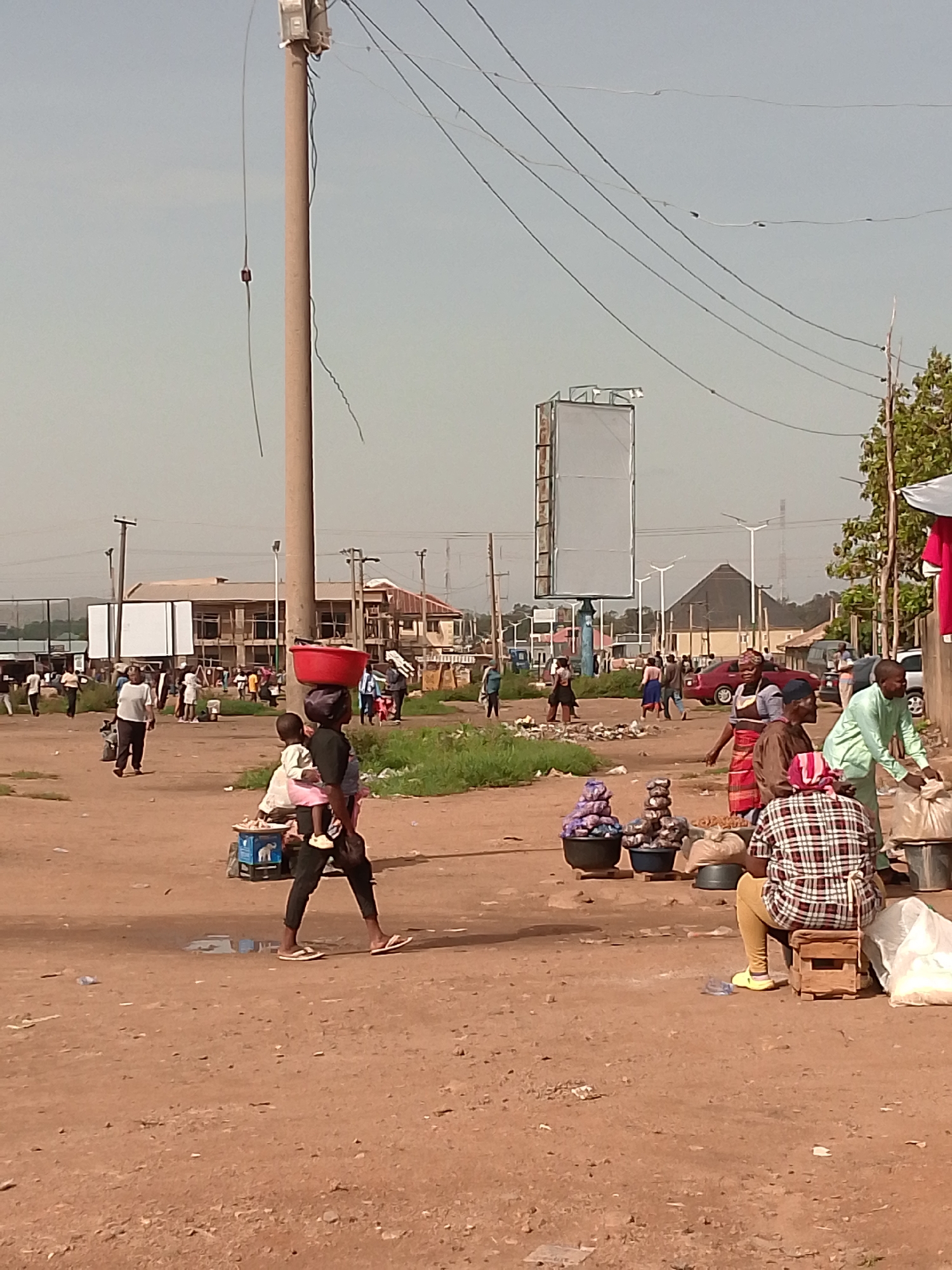 The image depicts an outdoor marketplace scene in Bukuru, Nigeria. In the foreground, a woman carries a red basin on her head and a child on her back, walking across a dirt path. Beside her, a child sits on a small cart. To the right, a person wearing a plaid shirt and yellow pants sits on a wooden stool, facing away from the camera, with bags of goods nearby. Further back, several other individuals are engaged in market activities, including a man in a green shirt handling a bag and a woman in a red and blue wrapper. Stalls with goods are visible, and various people are walking and interacting. The environment is dusty, with vegetation and buildings in the background. Overhead, power lines are strung across the sky, supported by a tall pole. A large, blank billboard is also visible in the distance. The overall impression is one of a busy, informal marketplace.