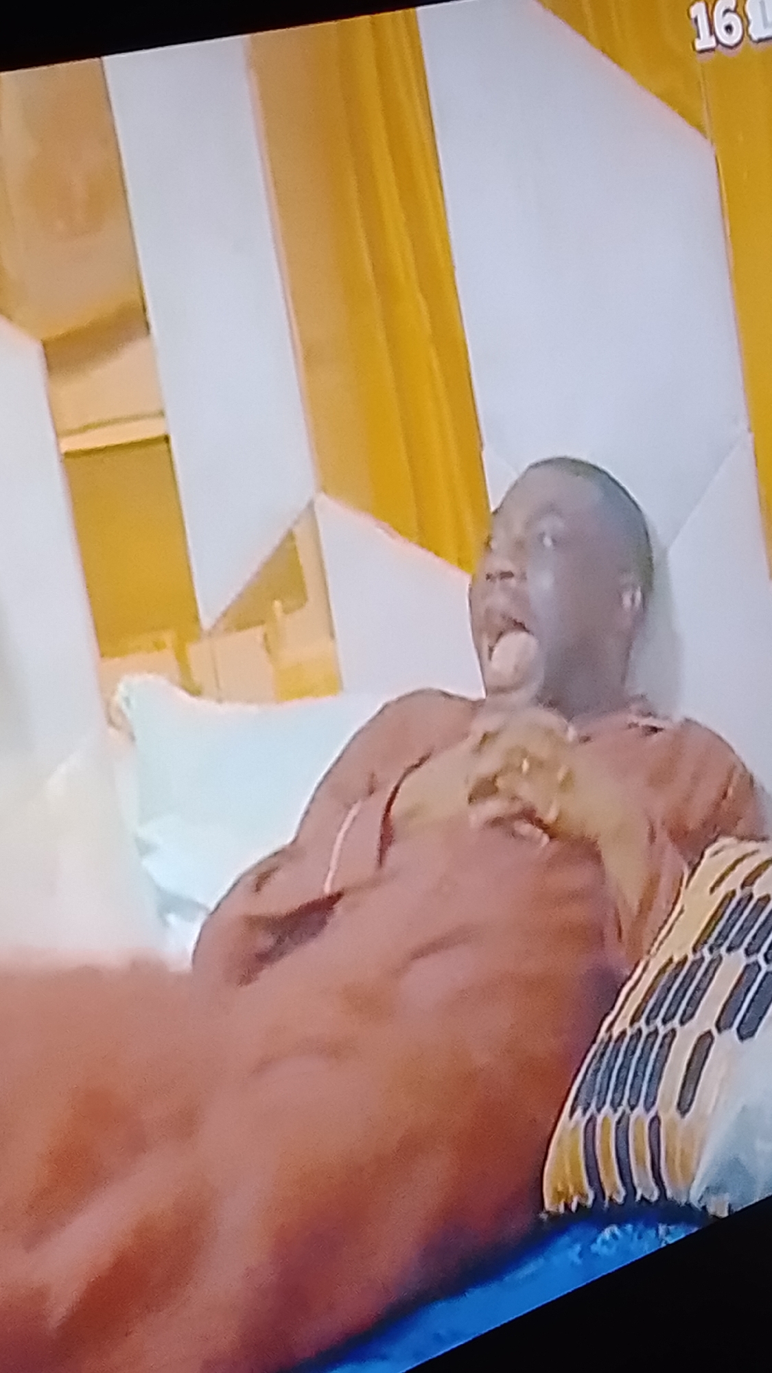 A person with dark skin is lying down in bed. They are wearing a brown garment that covers their body. Their hands are clasped together on their chest, and their mouth is open with their tongue visible. The background of the image consists of a wall with a geometric pattern of alternating white and yellow panels. There is also a pillow with a striped pattern visible on the right side of the image.