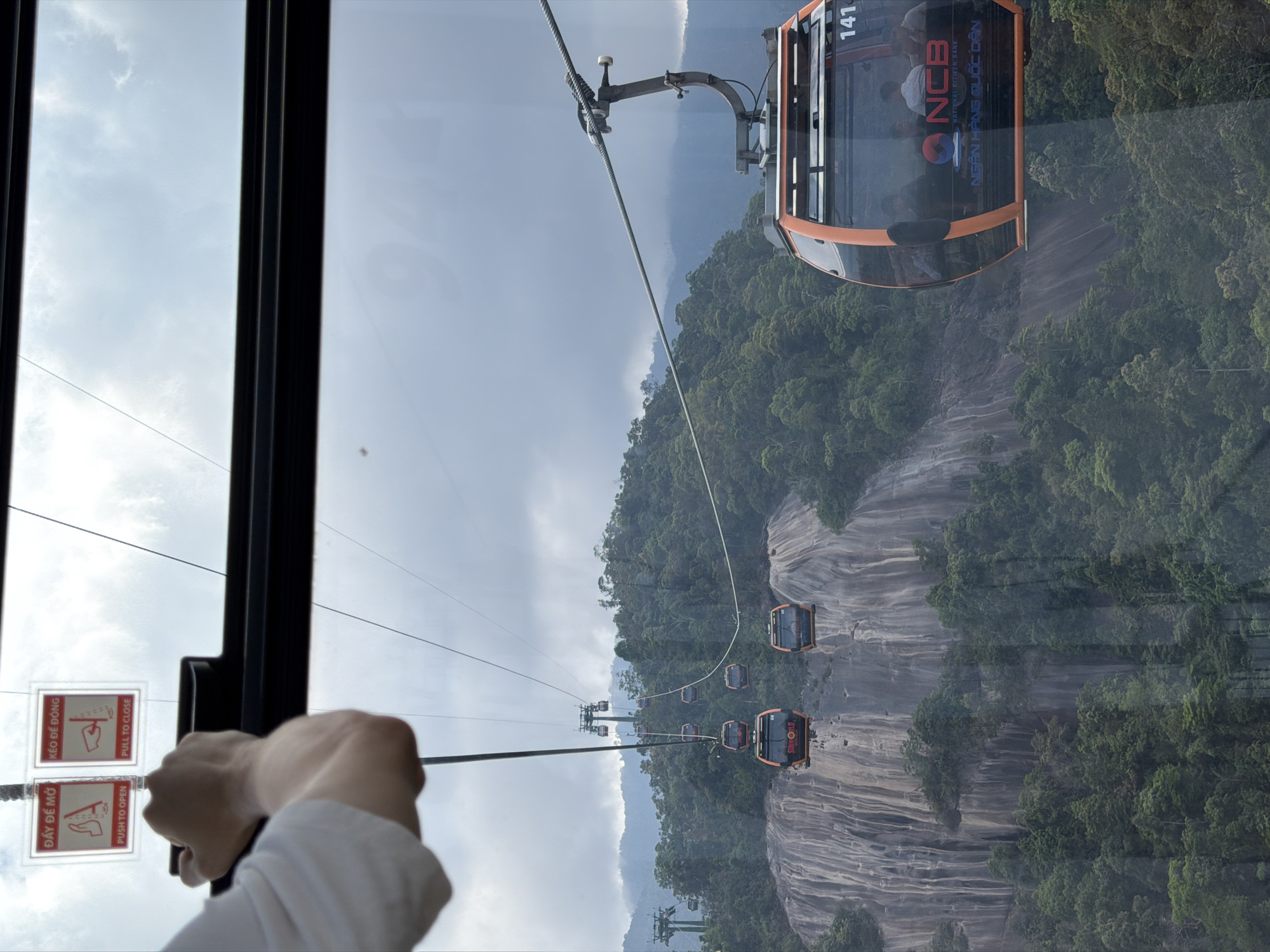 The photograph, taken inside a cable car, shows a view of the Ba Na Hills in Đà Nẵng, Vietnam. Several cable cars ascend along the line, traversing over a landscape of lush, green forest and distinctive rock formations. The sky is overcast with gray clouds. A person's hands are visible in the lower left, holding onto a black handle inside the cable car. Stickers with instructions in Vietnamese and English, "PUSH TO OPEN," and "PULL TO CLOSE," are affixed to the window. The number "141" and the "NCB" logo are visible on the closest cable car.