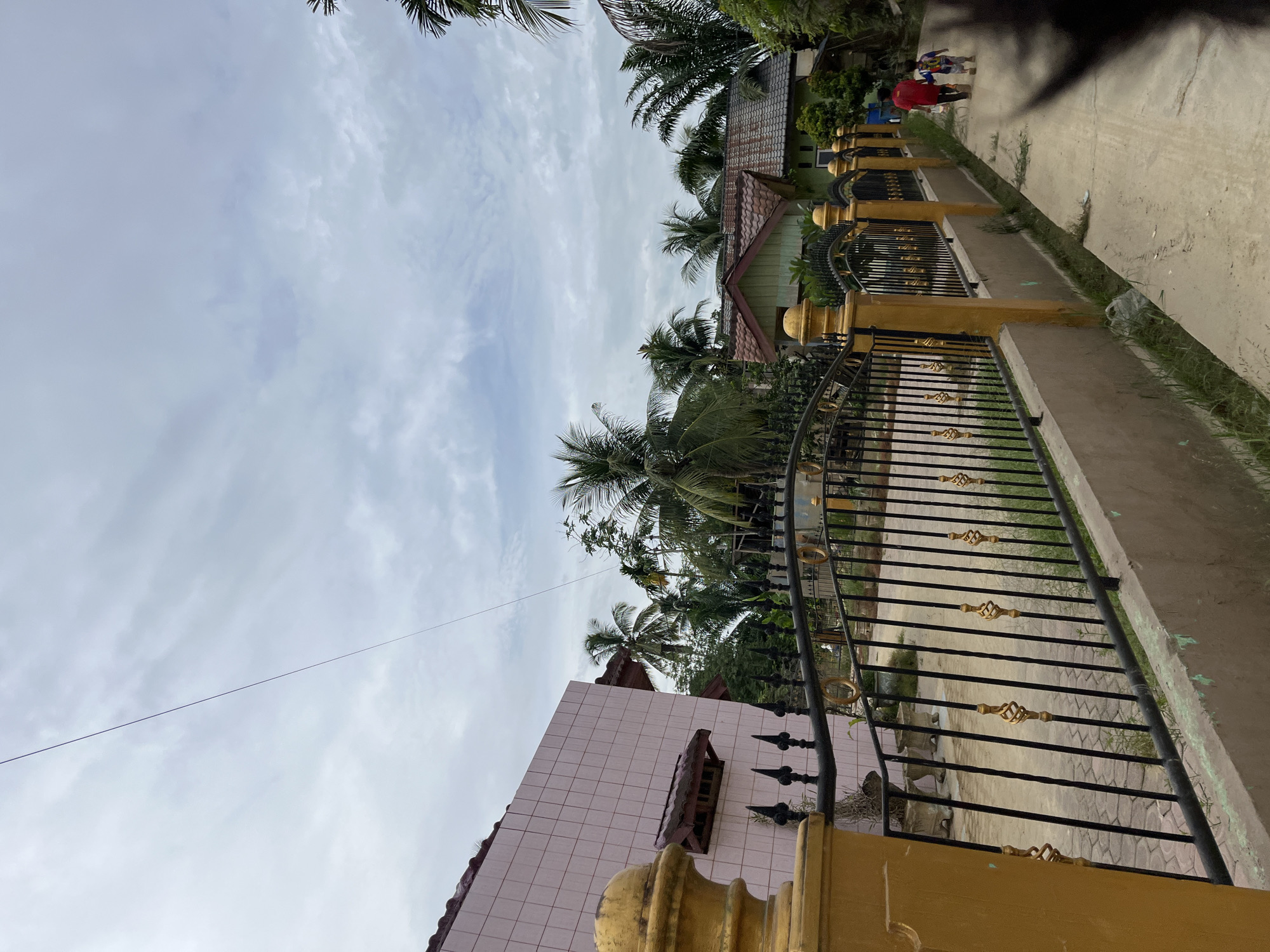 The image depicts a street scene in Menanggini, Indonesia, on an overcast day. In the foreground, a decorative yellow and black metal fence runs along a concrete pathway. To the left, a section of a light pink tiled building is visible, with a red-tiled awning over a small window. Beyond the fence, several houses are nestled amongst lush green palm trees and other tropical vegetation. Two people, a taller individual in a red shirt and a smaller child in a colorful shirt, are walking away from the camera down the pathway. The sky is filled with grey, diffused clouds.