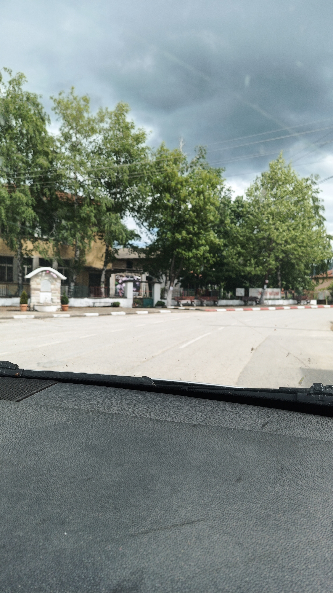 The view is from inside a car looking out onto a street in Gornoslav, Bulgaria. The dashboard and windshield are visible in the foreground. The street is straight and leads to an intersection. Buildings and trees line the street. The sky is cloudy and overcast. There are no people visible in the scene. A small, decorative stone structure stands near a building on the left side of the street. Red and white markings border the edge of the street.
