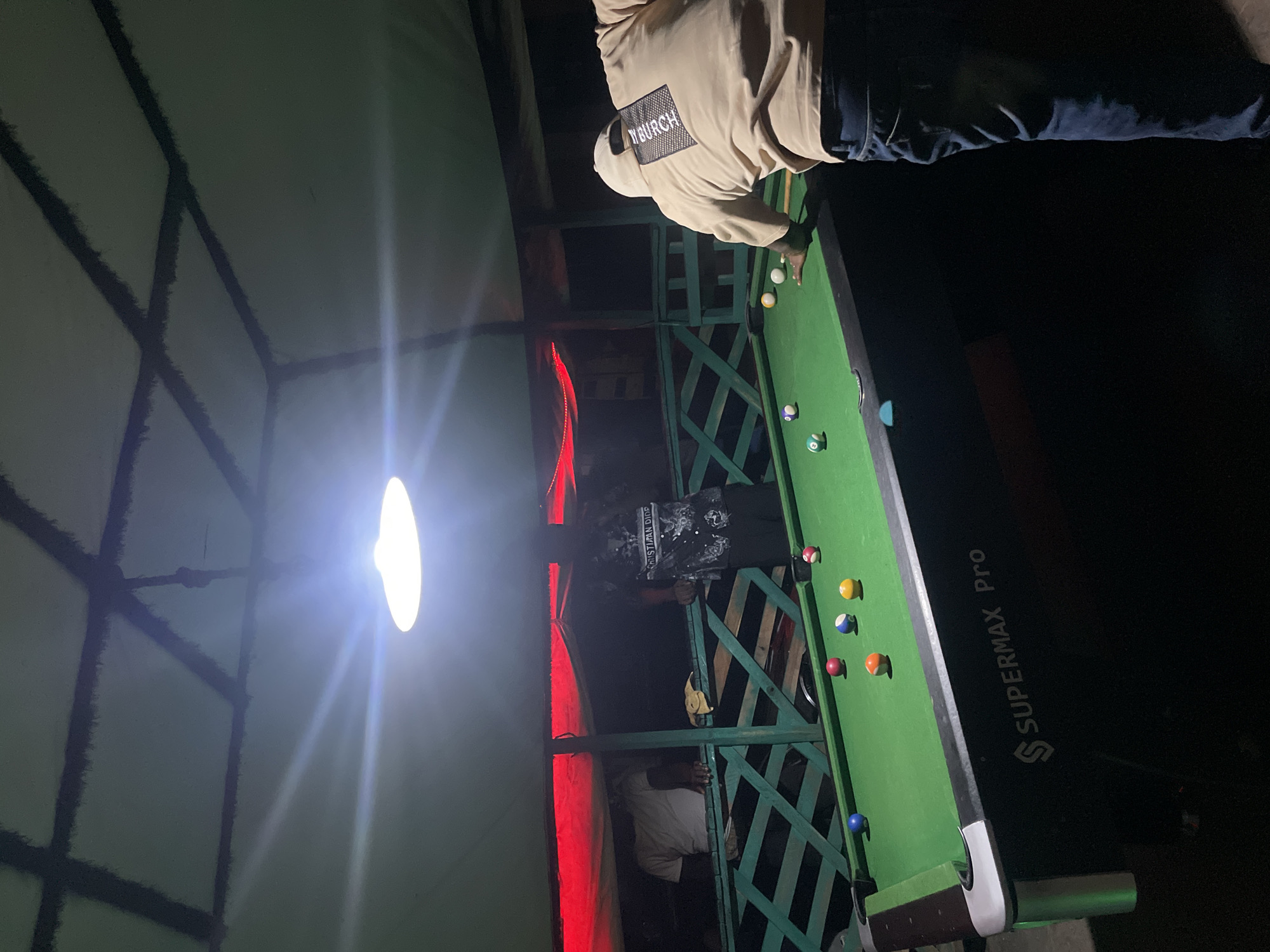 The image depicts a group of people playing pool at night. The pool table is positioned in an outdoor setting, possibly a veranda or a covered patio, which suggests an establishment in Lokoja, Nigeria.
One individual is seen preparing to take a shot with a cue stick. The pool table has several balls scattered across the green felt surface. The area is illuminated by an overhead light, causing a bright glare. The location features wooden fence, creating enclosure for the pool table. The word "SUPERMAX Pro" is visible on the side of the table.