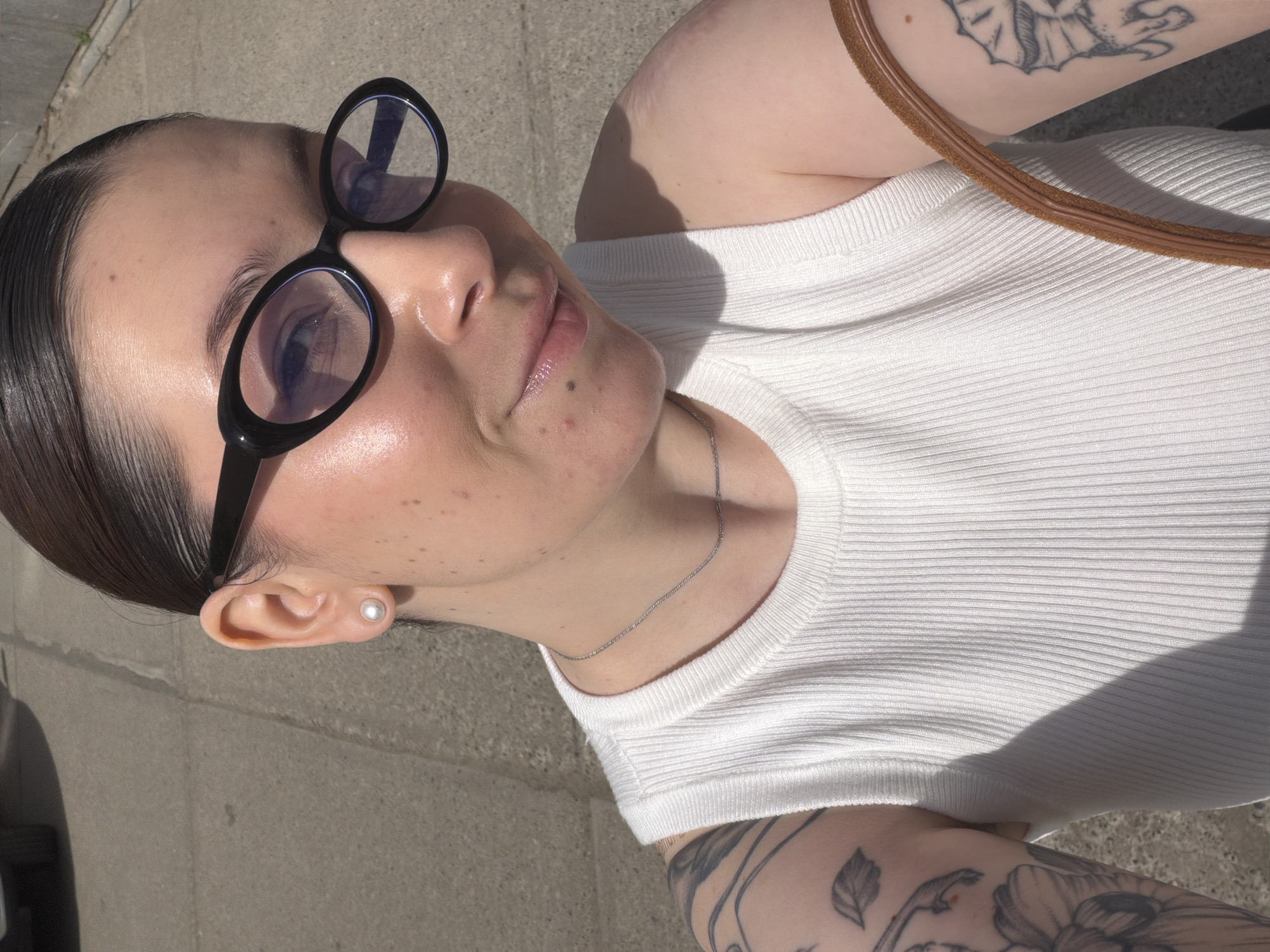 A woman with slicked-back dark hair is looking up at the camera. She is wearing round, black-rimmed glasses and a pearl earring in her left ear. She is wearing a white, ribbed tank top and a delicate silver necklace. Tattoos are visible on both arms. The background appears to be a concrete sidewalk. The location, Chaikovsky, Russia, is not definitively identifiable from the provided image.