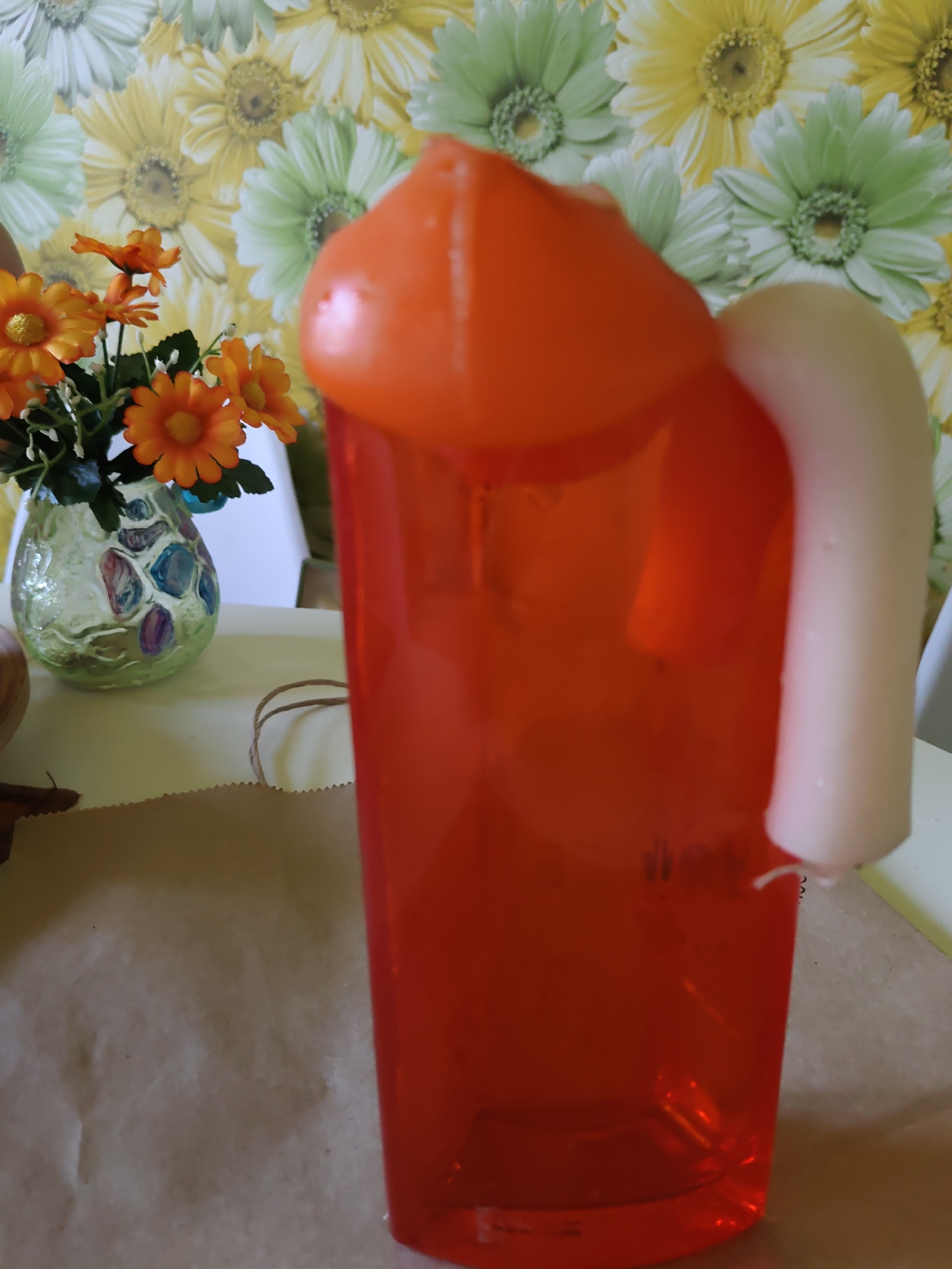 The image shows a close-up of a large, translucent orange plastic cup with a white handle, positioned in the foreground. The cup appears to be a novelty item, possibly a mug, with a rounded top resembling a lid or part of its structure. A white, curved handle is attached to the right side of the cup. Inside the cup, a red liquid is visible. To the left of the cup, a small bouquet of artificial orange flowers with yellow centers is placed in a green glass vase decorated with colorful abstract shapes. The vase is sitting on a light-colored surface. The background features a floral wallpaper with large, stylized flowers in shades of yellow, green, and white. A brown piece of paper or fabric is spread out in the lower part of the image, partially covered by the cup. The overall scene is brightly lit, with the focus primarily on the novelty cup and flowers. The image does not provide any information that would indicate its location in Rostov-on-Don, Russia.