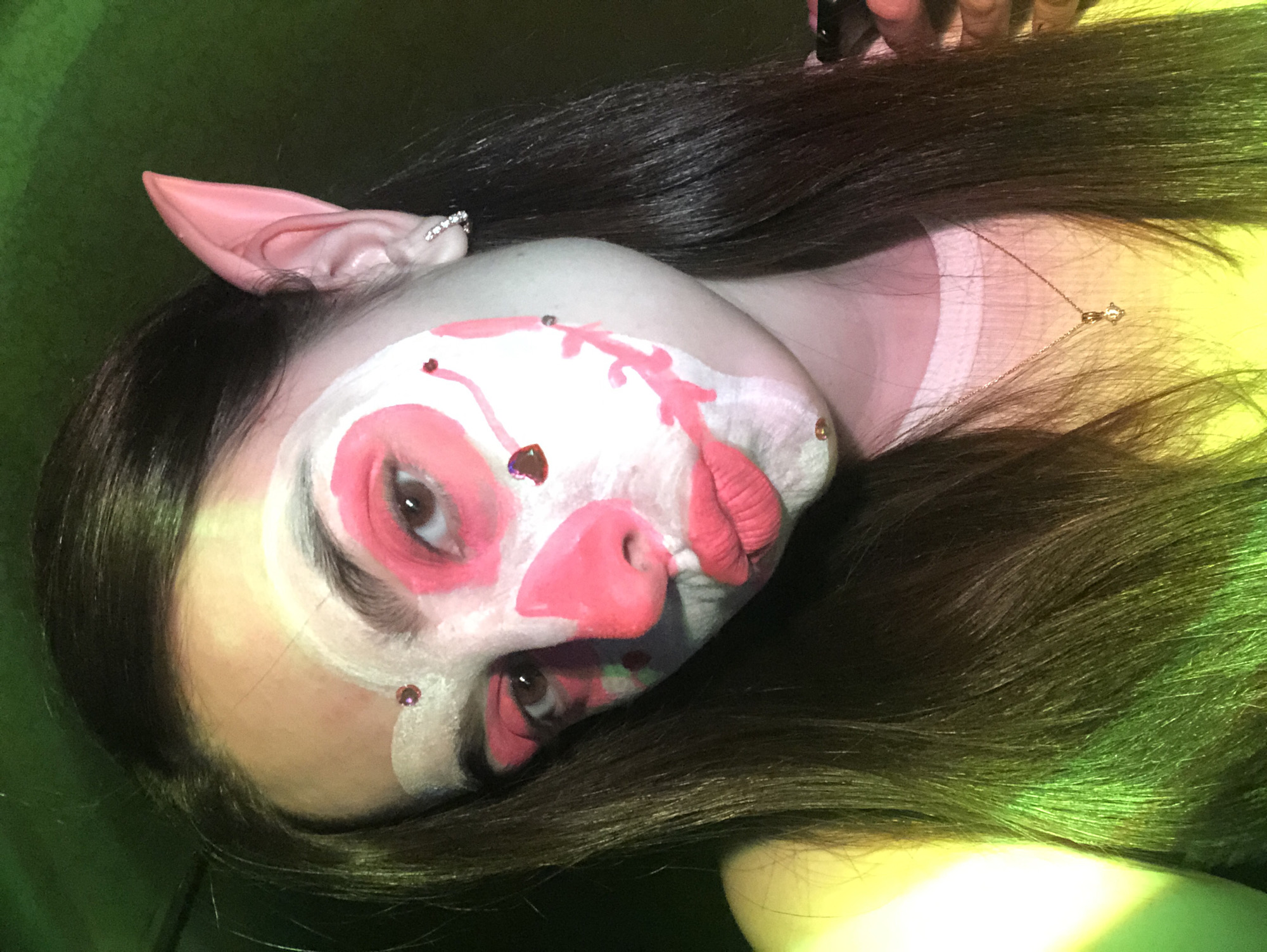 A person, likely female, with long, dark hair, is depicted in a close-up, slightly rotated portrait. She is wearing elaborate face paint that transforms her into a fantasy creature or pig-like character. Her face is covered in a white base, with prominent pink details defining her eyes, forming a stylized snout with visible nostrils, and creating textured, puckered lips. Small, reddish-pink rhinestones or gems are affixed to the white face paint, particularly above and below her eyes. She sports pink, pointed, prosthetic ears, resembling either elf or pig ears, with one ear adorned by a small, sparkling clip. A delicate gold-colored necklace with a tiny pendant is visible on her collarbone, partially obscured by her hair. Her long, dark hair is spread out around her head, catching green and yellow hues from the ambient lighting. The background is a dark, mottled green, with brighter yellowish-green areas, suggesting artificial or theatrical lighting. She looks directly at the camera with a neutral expression, showcasing her detailed makeup and accessories. The activity is a posed photograph. This scene is set in Noviy, Russia.