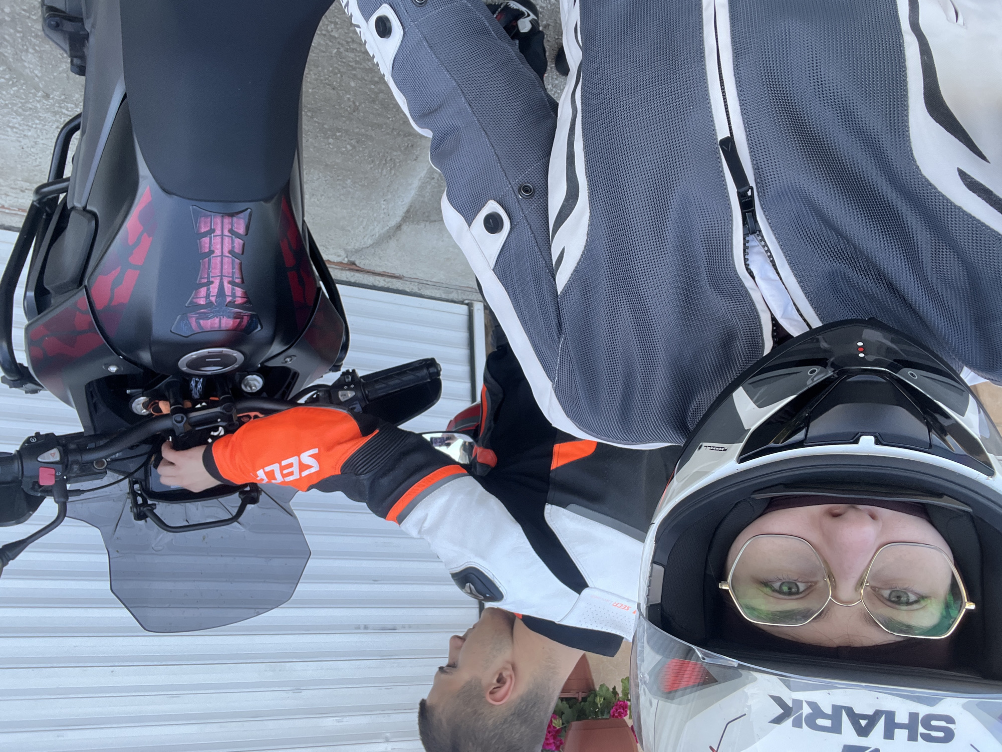 The image shows two individuals near a motorcycle in an unidentified city. The first individual, wearing a white and black motorcycle helmet and gold-rimmed glasses, is facing the camera. The second individual, dressed in a white and black motorcycle jacket with orange sleeves, is operating the controls of a black and red motorcycle. The setting appears to be an outdoor location near a garage or similar structure.