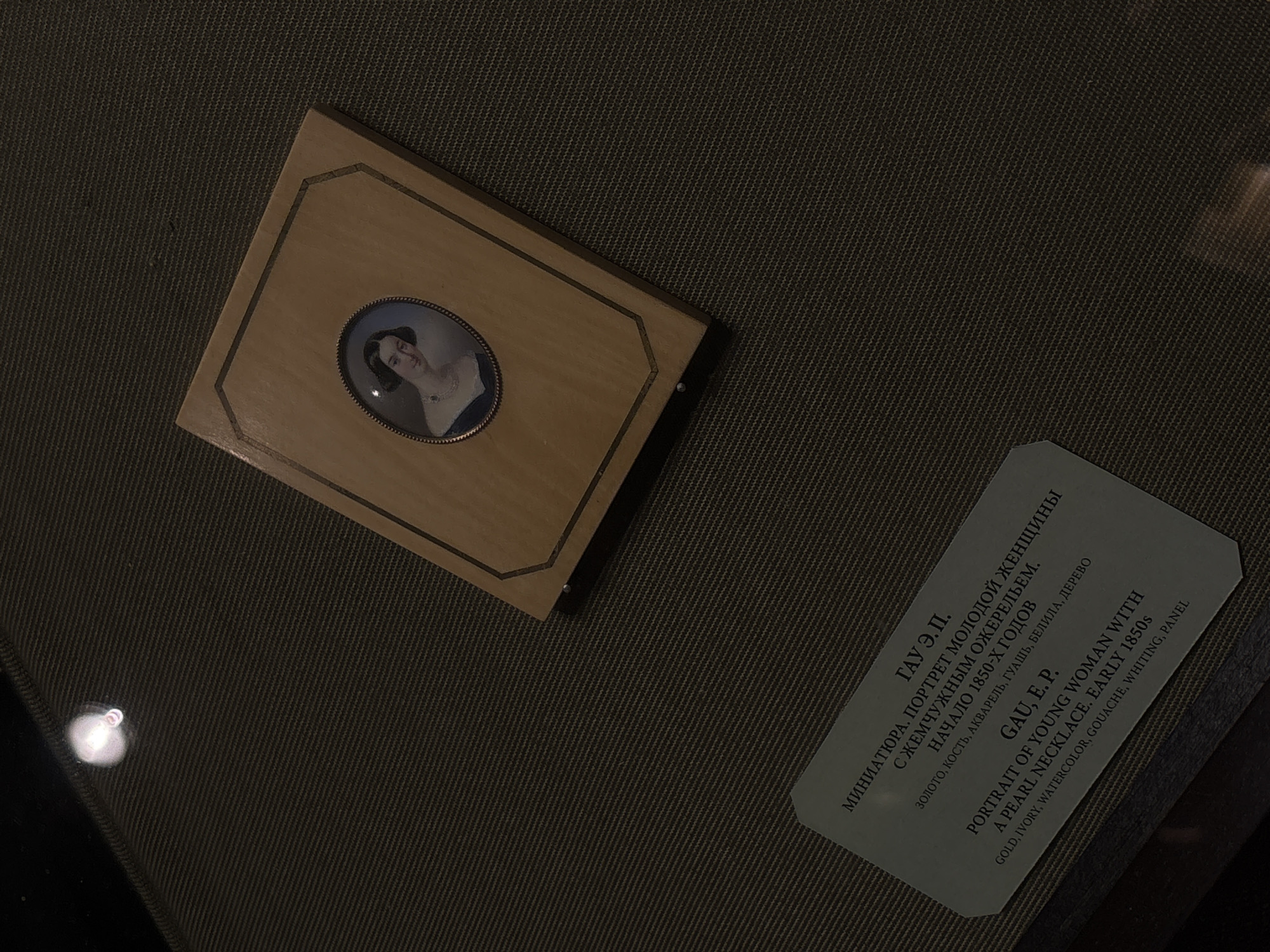 The image shows a framed miniature portrait of a young woman with a pearl necklace, dating from the early 1850s. The portrait is housed in a small, rectangular wooden case with a decorative border. To the right of the miniature is a label written in both Russian and English, identifying the artist as E.P. Gau and providing details about the artwork's materials (gold, ivory, watercolor, gouache, whiting, panel) and title ("Portrait of Young Woman with a Pearl Necklace"). The objects are displayed on a dark brown, textured surface, likely within a museum or gallery setting, possibly in Moscow, Russia, given the presence of Russian text on the label.