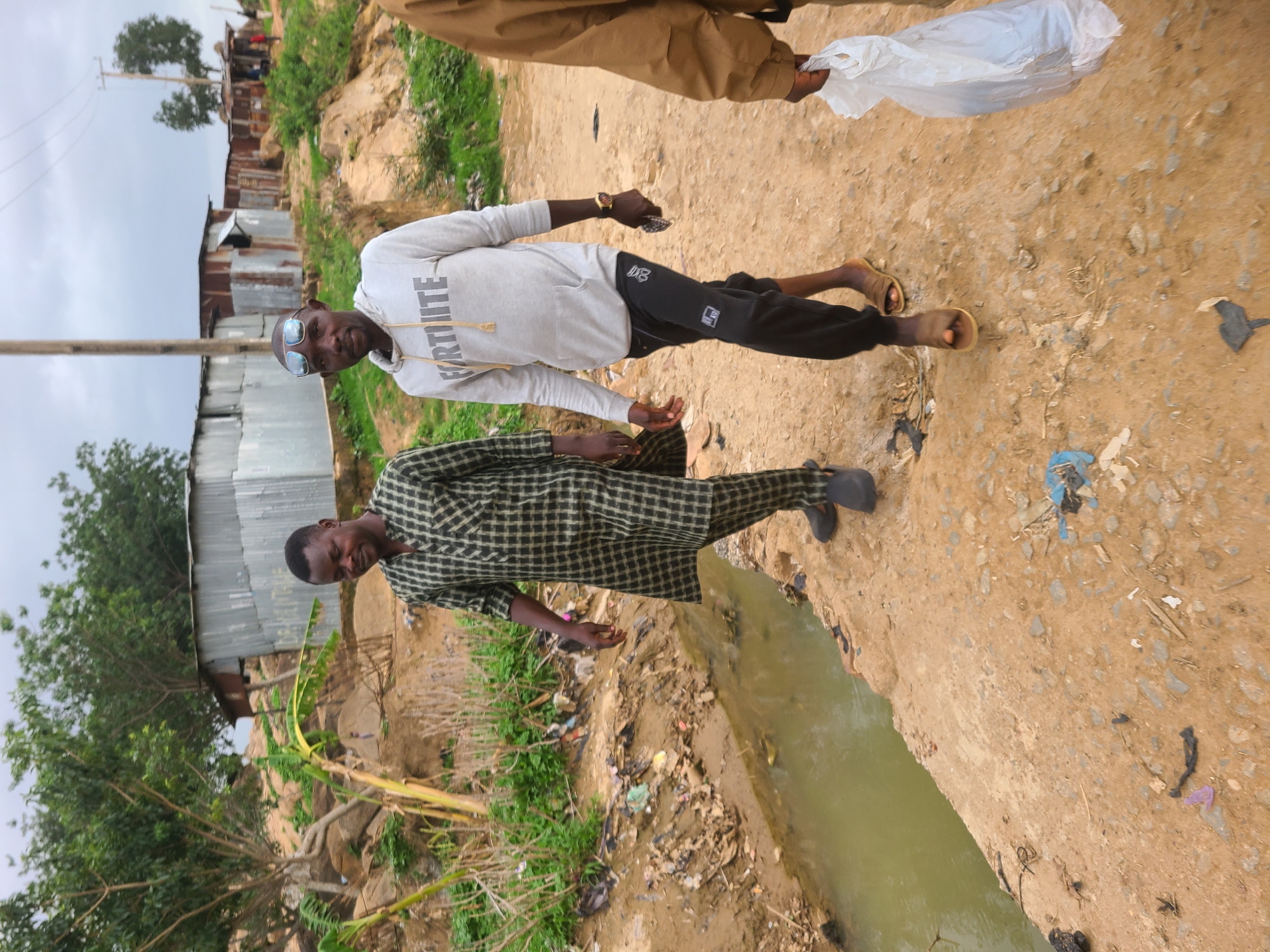 Two men are standing on a dirt road beside a narrow canal of murky water. Debris and vegetation litter the canal bank. One man is wearing a checkered robe and black shoes. The other man is wearing a light gray sweatshirt with the word "FORTNITE" printed on it, black pants, sunglasses, and sandals. A third person's legs and hand holding a white plastic bag are visible in the upper right corner. Buildings and trees are visible in the background.