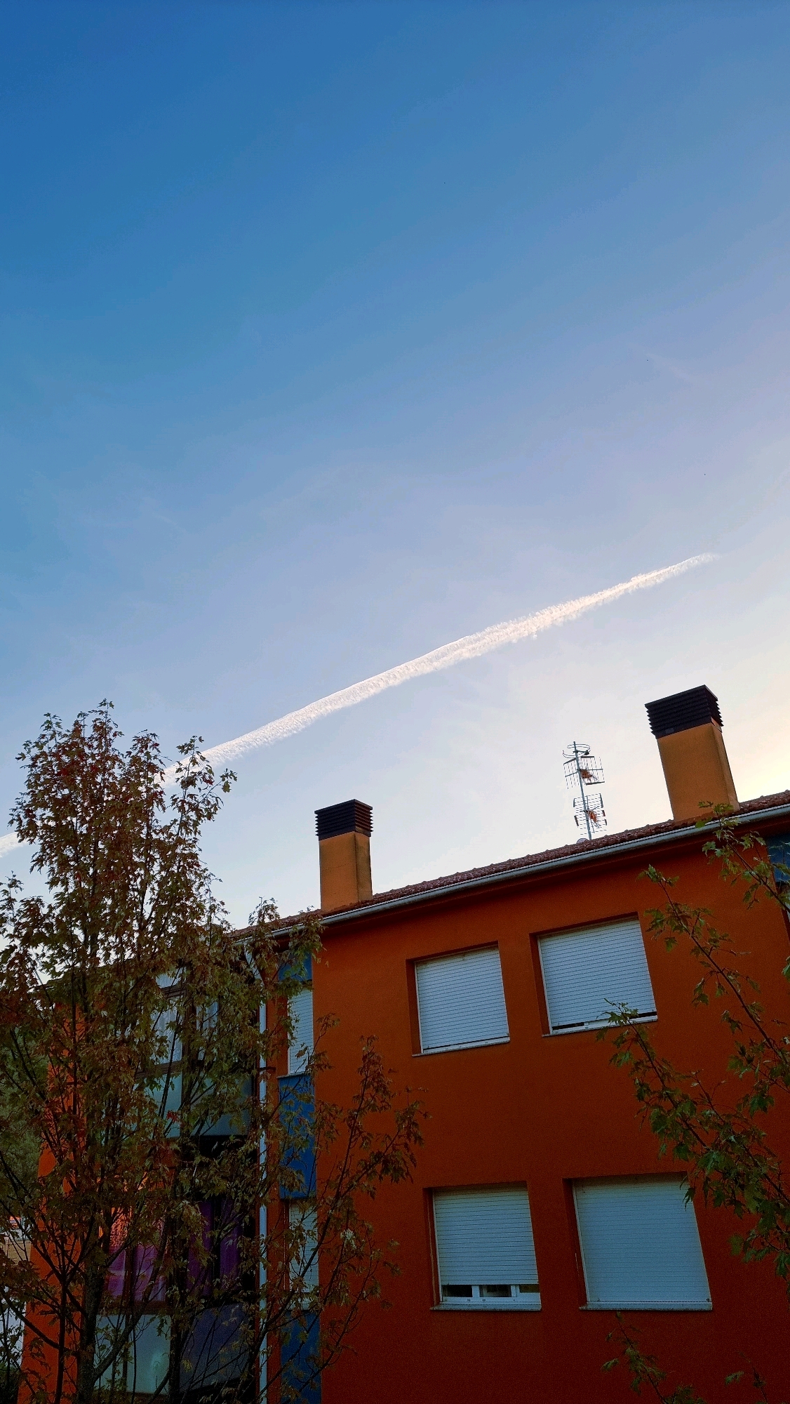 An upward-angled shot captures a bright orange building in Sant Joan Les Fonts, Spain, against a gradient blue sky. A thin, white contrail stretches diagonally across the upper portion of the sky. The building has a red-tiled roof and features several windows with closed white shutters. Two dark-capped chimneys rise from the roof. A television antenna is positioned between the chimneys. To the left of the building, a tree with autumnal foliage, displaying hues of brown, red, and green, partially obscures the structure's left side.