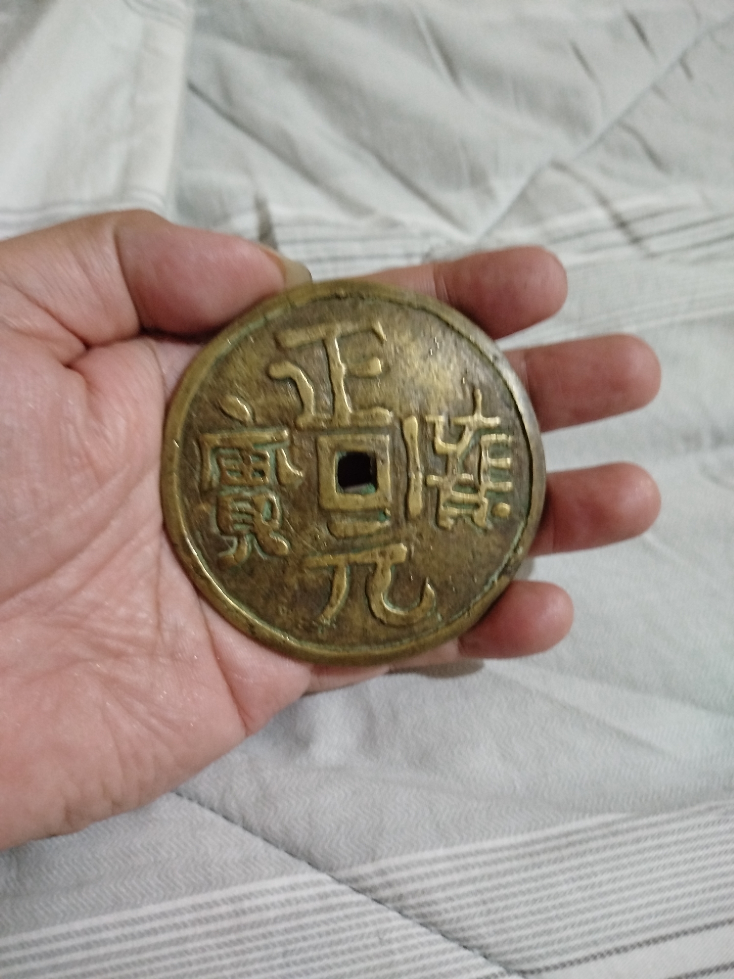 A close-up shot shows a hand holding a round, brass-colored coin with a square hole in the center. The coin has Chinese characters inscribed on it. The background is a light-colored, textured surface, possibly a bedsheet. The image was taken in Bandungrejo, Indonesia.