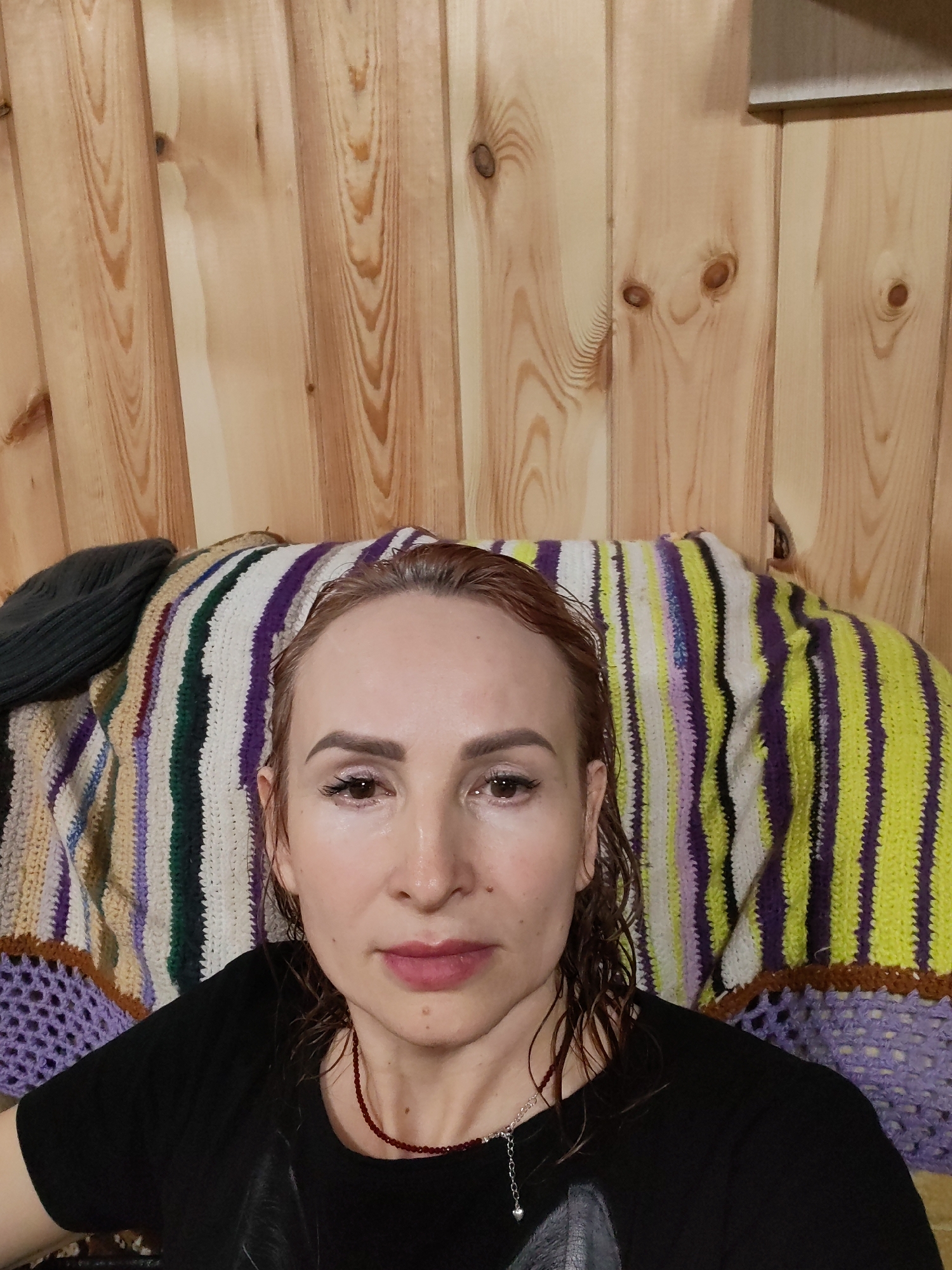 A medium close-up shows a fair-skinned woman with reddish-brown, damp hair. She is wearing a black t-shirt and a necklace with red beads and a silver pendant. Her expression is neutral. She appears to be sitting or leaning against a multi-colored striped knitted blanket or throw. Behind her, the background consists of a wooden wall made of vertical planks. The image was taken indoors, likely in a private residence in the city of Chaikovsky, Russia.