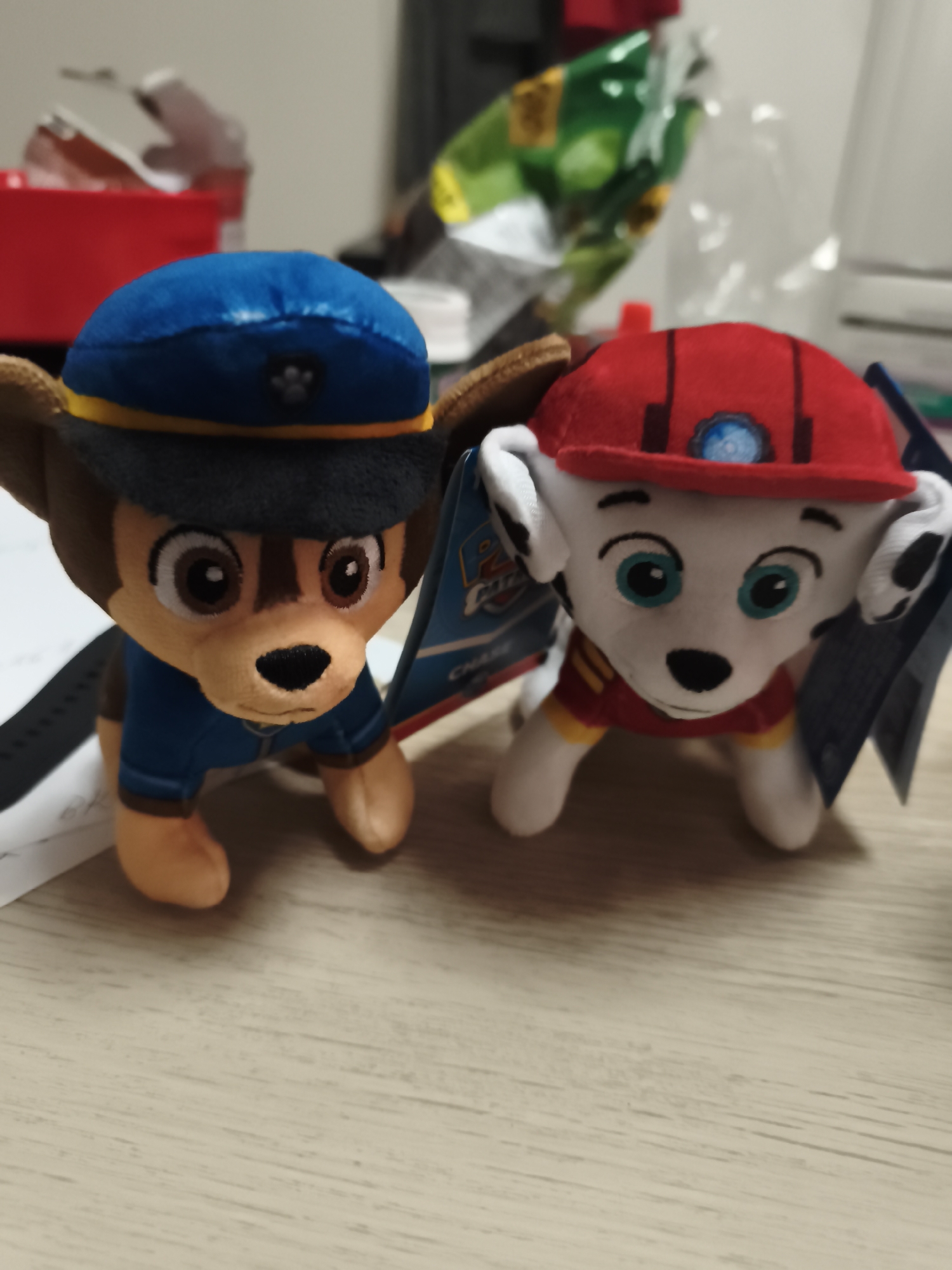 This indoor photograph features two plush toy puppies from the animated series "Paw Patrol," placed on a light brown, wood-grained surface. The toys are in sharp focus, while the background is blurred.
On the left, a brown-furred puppy plush, identifiable as Chase, wears a blue police uniform and a matching blue hat with a yellow band and a dark grey paw print emblem. Its eyes are large and brown. An attached retail tag is partially visible beside it, with the word "CHASE" discernible.
To the right, a white-furred puppy plush with black spots, identifiable as Marshall, is dressed in a red firefighter uniform and helmet. The red helmet has a central blue circular emblem with a white design. This toy has prominent light blue eyes. Another retail tag is visible next to it.
The setting appears to be a casual indoor environment, possibly a desk or table, under artificial lighting. In the soft-focused background, a red container or box is visible to the left, and further back, there are indistinct items including some crinkled green and transparent plastic packaging. The image was taken in Huntingdonshire, United Kingdom.