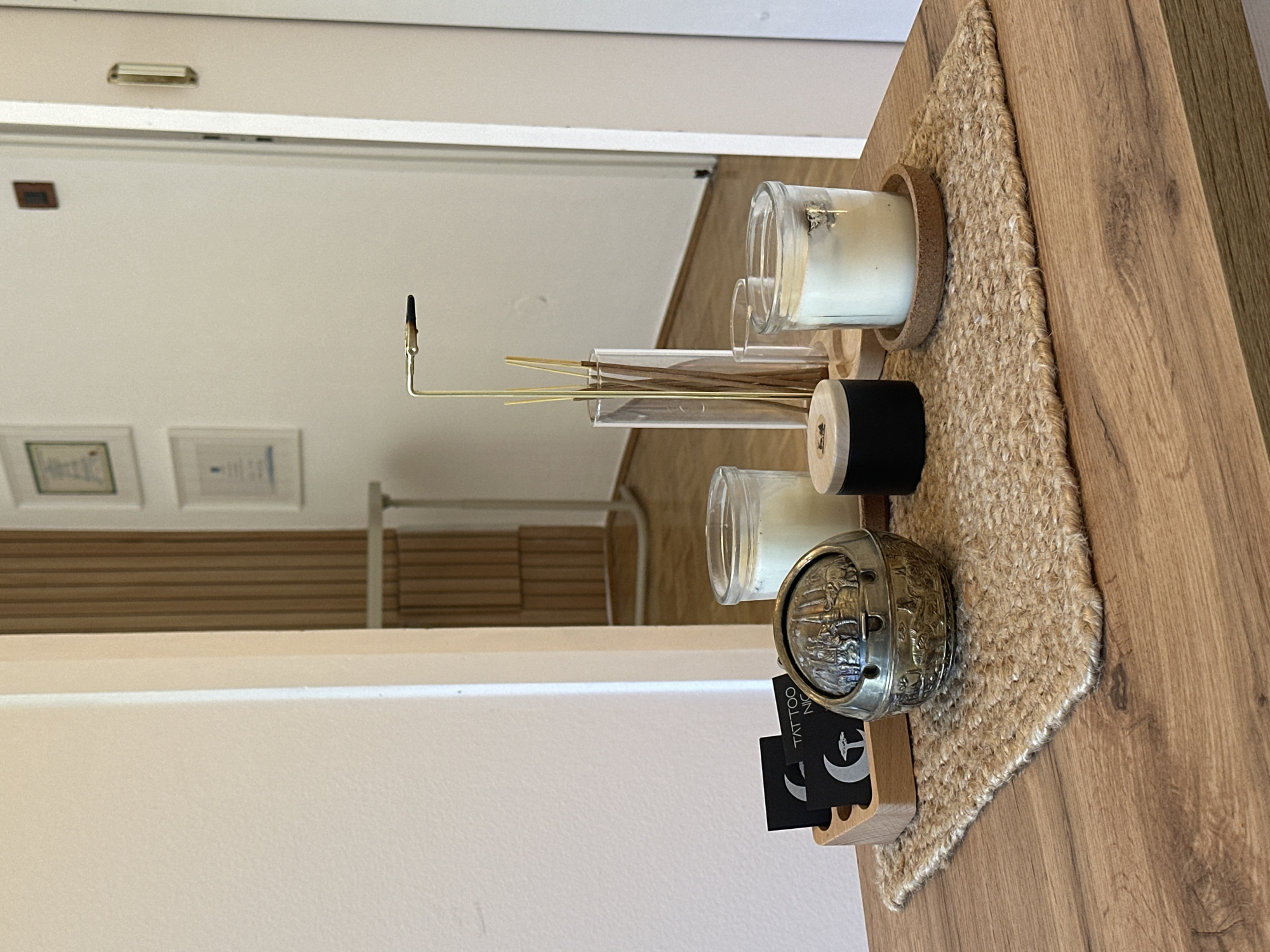 The image, captured in the City of Zagreb, Croatia, displays a meticulously arranged collection of objects on a light-colored wooden table. A textured, woven mat made of light-brown natural fibers forms the base for the arrangement.
From left to right on the mat, a slender, golden L-shaped incense holder supports a lit incense stick, emitting a thin wisp of smoke. Behind it, a clear cylindrical glass container holds several unlit incense sticks. Two clear glass jars, each containing white candle wax, are prominent; the jar on the far right rests on a cork ring coaster. Between these jars, a distinct cylindrical object made of black and natural wood is visible. Further to the right, an ornate metallic sphere, possibly a decorative ash tray or container, sits on the mat. Beside it, a small wooden block holds a stack of black business cards.
The background shows a reflection of an interior space, likely a room or hallway. White walls are visible, and on one wall, two framed documents or certificates are displayed, though their text is illegible. Below these frames, a section of horizontal wooden slatted paneling can be seen. The bright, even lighting suggests it is daytime.
No people are present in the image. The visible text includes "TATTOO" and "NIC" on the black business cards, accompanied by a logo featuring a crescent moon and a stylized symbol that resembles an epsilon or a part of a euro sign. The overall scene conveys a sense of tranquility and order, indicative of a professional or personal setting designed for comfort.