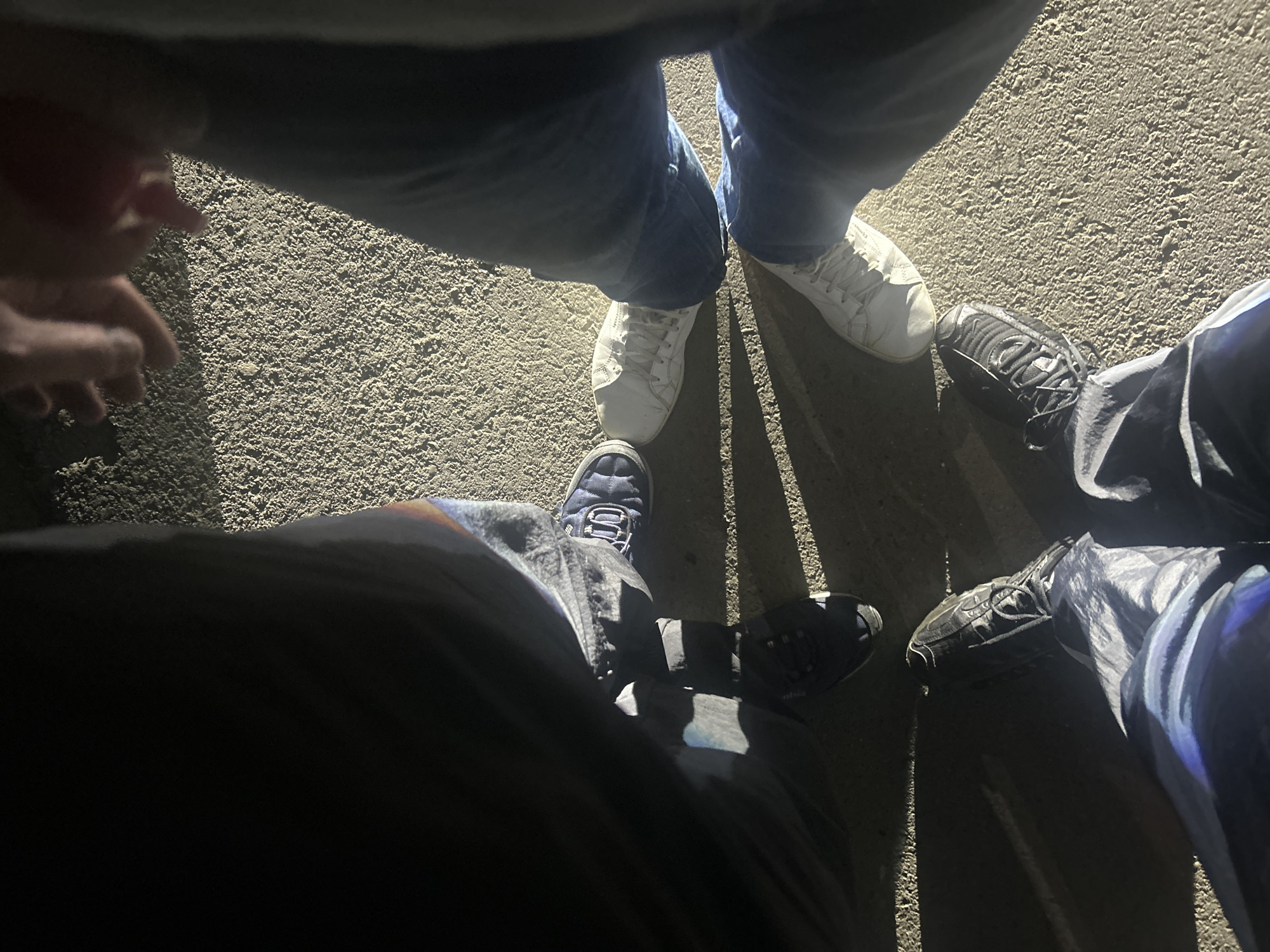 Four people stand in a circle on an asphalt surface. Each person is wearing shoes. The first person is wearing white sneakers and blue jeans. The second person is wearing dark running shoes and dark pants. The third person is wearing white sneakers and blue jeans. The fourth person is wearing quilted blue sneakers, pants are not entirely visible. A hand is visible on the left side of the image. The location and details about Chaikovsky, Russia are unknown.
