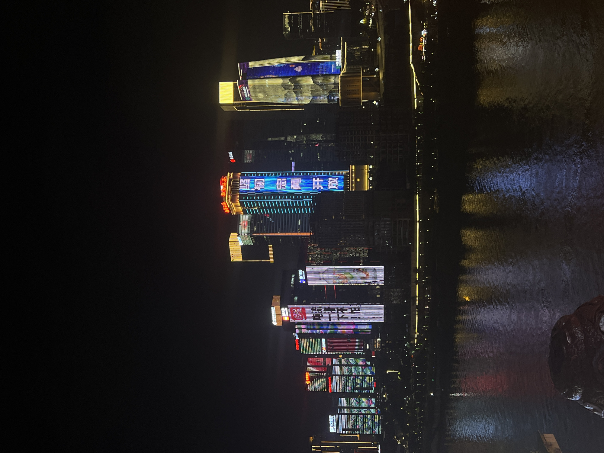 The image depicts a nighttime cityscape in the Yuzhong District, China, characterized by illuminated skyscrapers and their reflections on a body of water. The buildings are adorned with vibrant, colorful LED displays, showcasing various patterns, Chinese characters, and imagery. One prominent skyscraper on the right features a vertical display with blue and white patterns resembling mountains and water. To its left, another building displays bright blue vertical text. Other buildings in the skyline are lit with a mix of red, green, yellow, and white lights, some with intricate geometric designs. The foreground of the image shows the dark surface of the water, upon which the lights from the city are reflected, creating shimmering streaks of color. The overall atmosphere is one of urban dynamism and nighttime illumination.