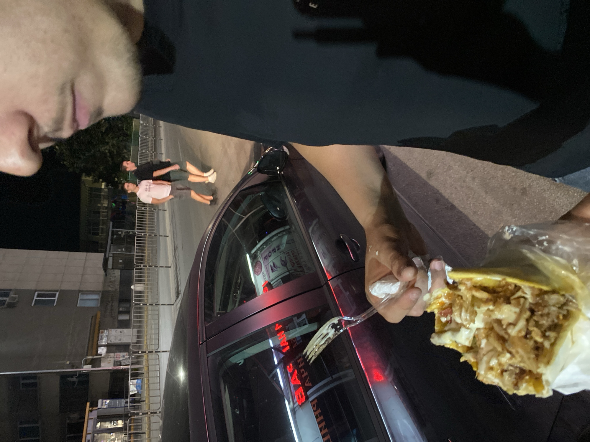 In an urban environment at night, an individual is holding a wrapped food item, possibly a gyro or shawarma, with a plastic fork and napkin. They are positioned in front of a dark-colored sedan. In the background, two young men are walking away from the camera on a sidewalk bordered by a metal fence and a building. The scene appears to be on a street in the city of Unknown, Unknown.