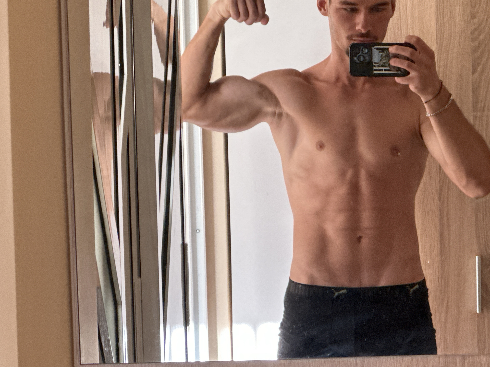 A shirtless young adult male, with tanned skin and defined abdominal muscles, poses in front of a mirror. He is flexing his left bicep and holding a smartphone in his right hand, taking a selfie. He is wearing dark, possibly athletic, shorts. The background consists of a wall with wood paneling and a large, multi-paneled mirror that reflects the scene and the surrounding environment, which appears to be an indoor space. The lighting suggests it is daytime. There are no discernible elements that place this scene specifically in Varna, Bulgaria.