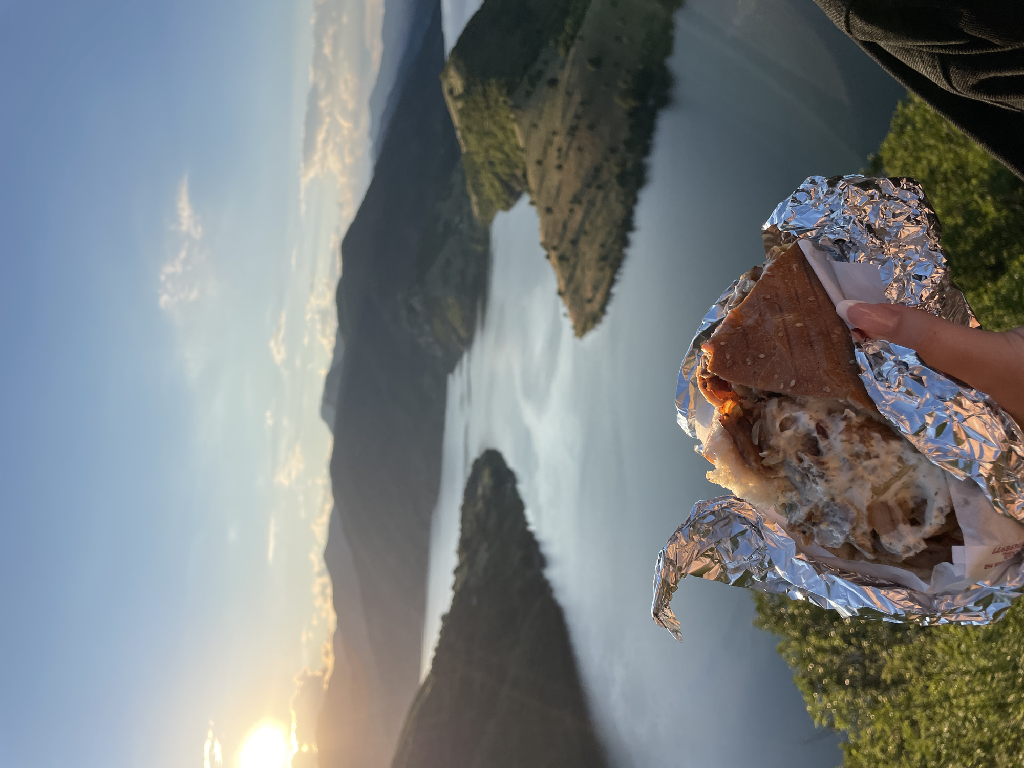In the foreground, a hand with manicured nails holds a partially unwrapped sandwich filled with meat and sauce. The sandwich is encased in reflective foil. Behind the sandwich, a panoramic view unfolds of a large, winding body of water, likely a reservoir, surrounded by numerous rolling, tree-covered hills and mountains. The sun is setting, casting a warm glow on the scene and creating a dramatic interplay of light and shadow across the landscape. The sky is a gradient of blue with scattered clouds catching the sunlight. This scene is located near the city of Borovitsa, Bulgaria, specifically overlooking the Borovitsa Reservoir.