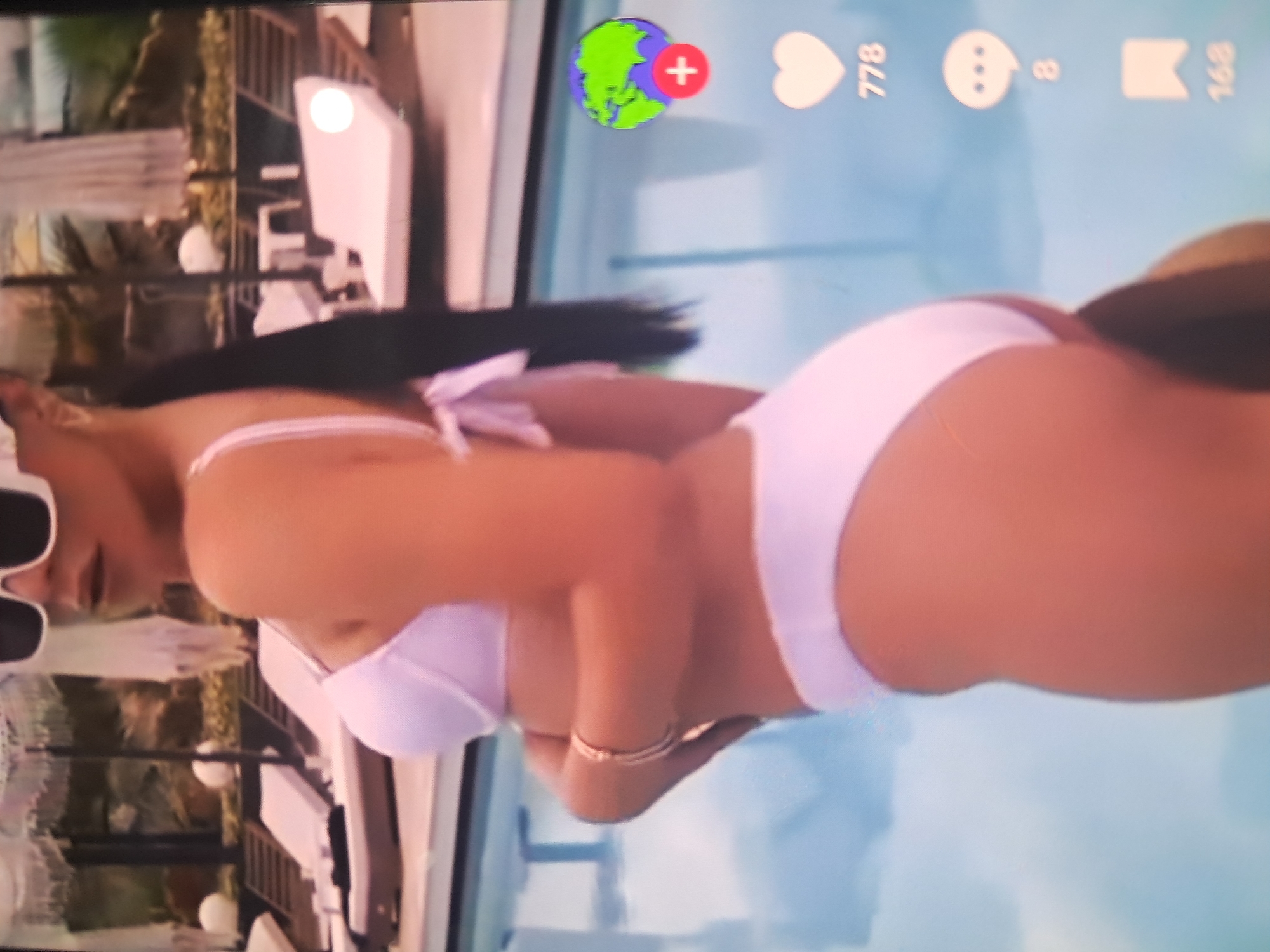 The image displays a poolside scene, vertically oriented, appearing to be a screen capture of a horizontally displayed video or photograph. The primary subject is a female individual lying on her back, wearing a white two-piece bikini and white-framed sunglasses. Her left arm is bent, resting near her abdomen, adorned with a thin bracelet. Her right arm extends towards her head, partially obscuring dark hair.
The background consists of a light blue swimming pool on the left, reflecting the sky. To the right of the subject, several white lounge chairs with dark cushions are arranged. Behind the chairs, architectural elements of a building are visible, featuring railings and multiple white spherical lights. Lush green vegetation, including palm-like trees, completes the background.
On-screen overlay elements are visible along the bottom-left edge of the image (in its current vertical orientation), including the numbers "168", "8", and "778", alongside a bookmark icon, a speech bubble icon, a heart icon, and a globe icon with a red plus sign. This scene is located in Blagoevgrad, Bulgaria.