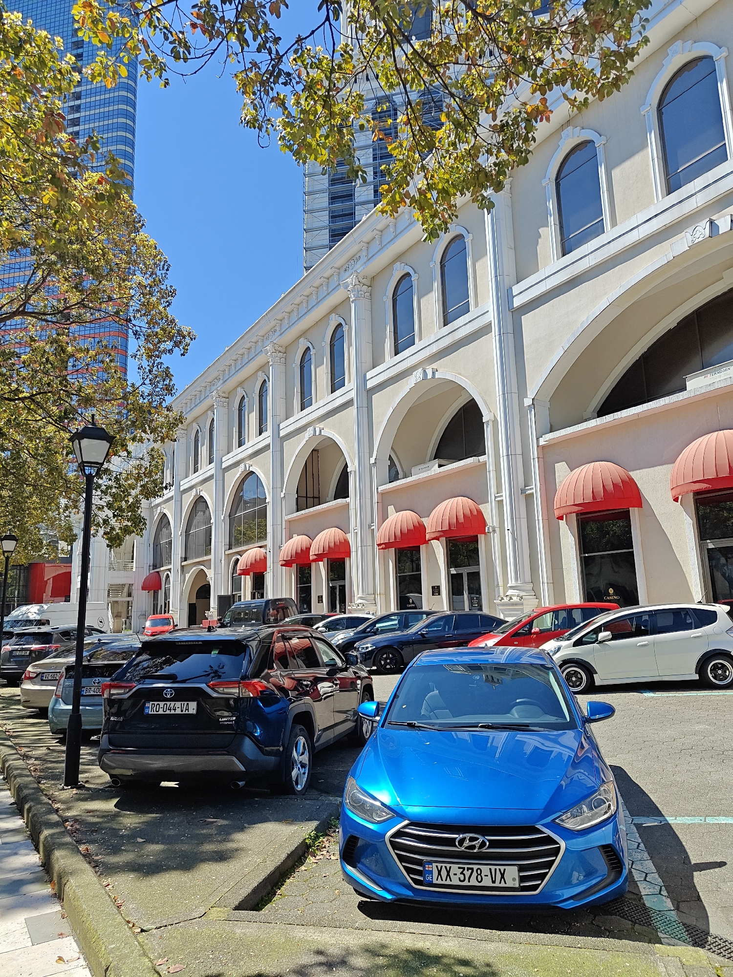 The image captures a street scene in Batumi, Georgia. In the foreground, a bright blue Hyundai sedan with the license plate "XX-378-VX" is parked on a section of the street with visible pavement markings. Directly behind it is a black Toyota RAV4 with license plate "RO-044-VA," parked near a lamppost. Several other cars are parked behind these two, including a white hatchback and a red car.
Behind the cars, a large, off-white building dominates the scene. The building's facade features a series of arched windows and entrances with red awnings. The word "CASINO" is visible above one of the entrances. The building’s design includes decorative architectural elements such as pilasters and cornices.
Above the building, a tall, modern skyscraper with a glass facade can be seen. The scene is bathed in sunlight, casting shadows from the cars and trees. Branches with yellowing leaves are visible in the upper left corner of the image, suggesting that it is autumn. The sky is clear and blue.