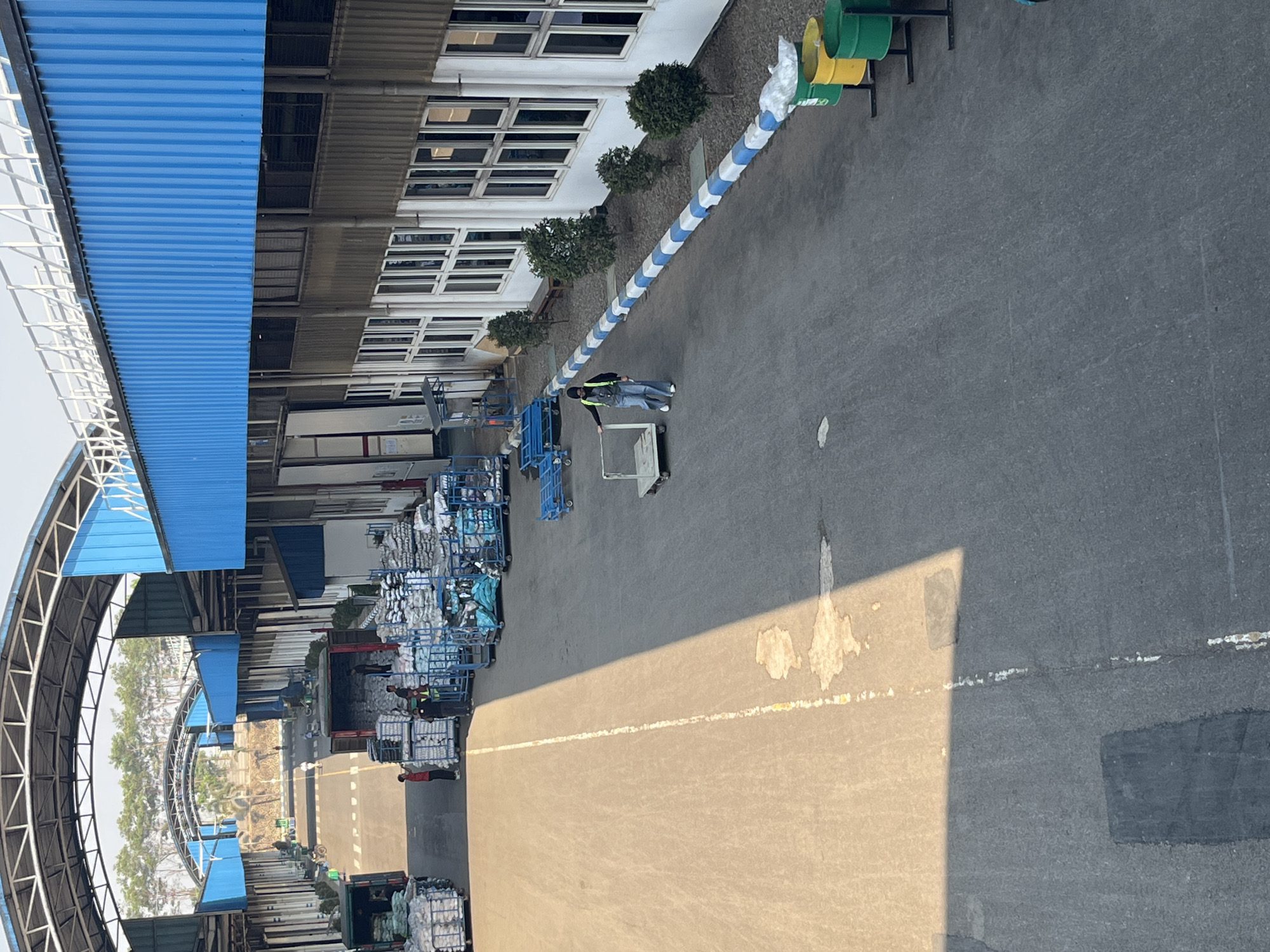 This image shows an exterior view of a warehouse or factory area in Pelang, Indonesia. The scene depicts a large, paved outdoor space with a covered walkway or tunnel structure extending overhead, supported by metal beams. On the right side of the image is a long building with a blue corrugated metal roof and numerous white-framed windows. A person wearing a yellow vest and blue pants is seen pushing a trolley along the paved area, adjacent to a low wall decorated with blue and white alternating blocks. Further down the paved area, several green and yellow bins are visible. On the left side of the image, multiple trucks are parked, some with their rear doors open, revealing stacks of goods wrapped in plastic. Several people, some in red shirts and others in vests, are present near the trucks and piles of goods. The ground is asphalt, with a yellow line marking a lane and patches of discoloration and what appears to be dried liquid. Trees are visible in the background beyond the covered structures. The overall impression is a busy industrial or logistics hub.