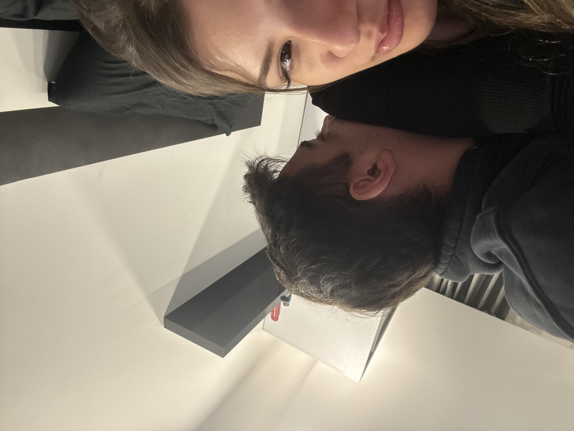 A selfie-style shot shows a woman and a man lying down. The woman is facing the camera with light brown hair, and her lips are visible. The man is positioned behind her, and only his head is visible. He has dark hair and is wearing a dark-colored hoodie. The background appears to be a room with white walls. In the lower part of the image, there is a shelf in shades of gray and white. The picture has been taken from an unusual angle. Sofia, Bulgaria is not discernible from the given context.