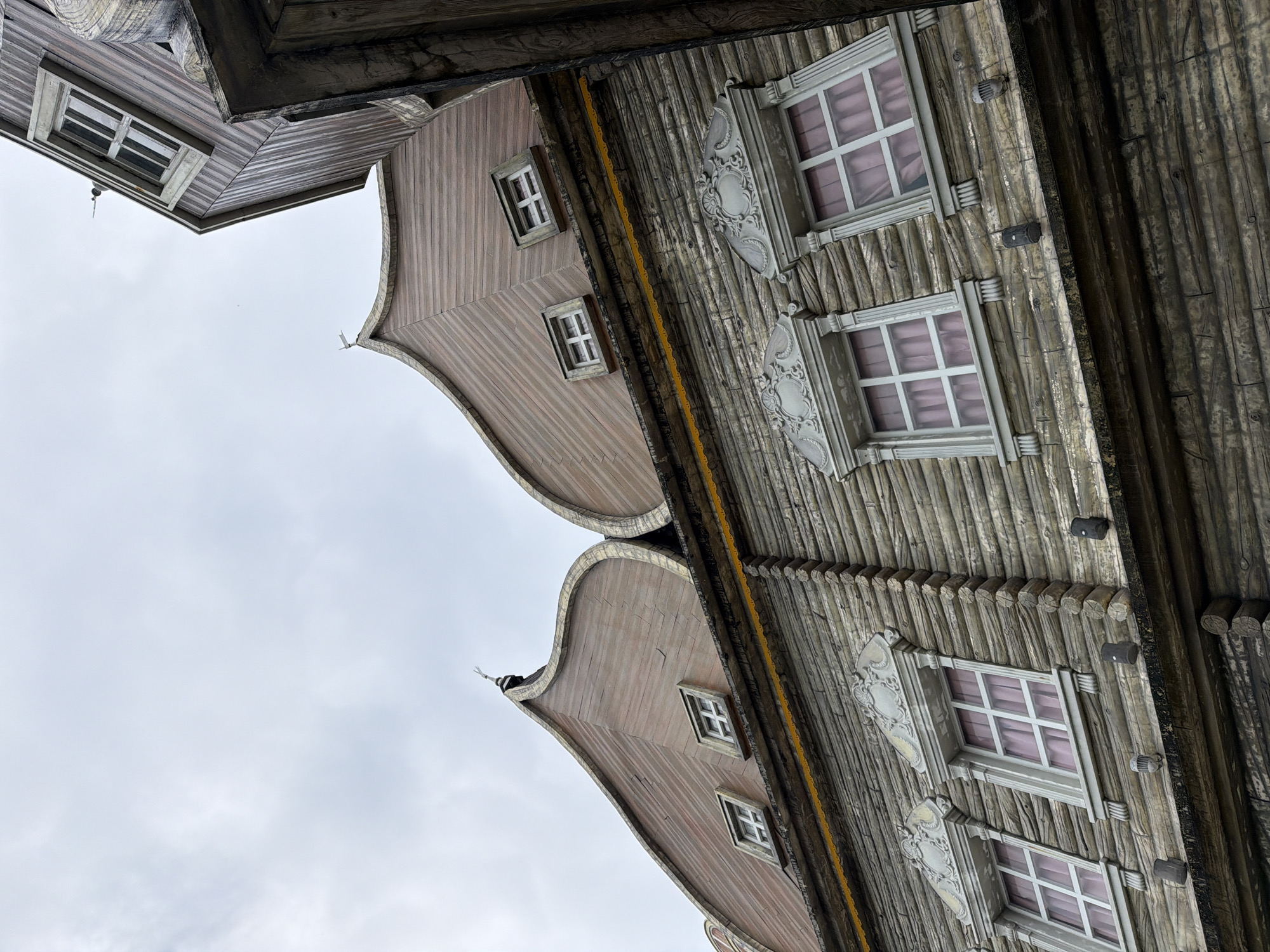The image shows a low-angle exterior shot of a building's facade, likely at an amusement park or themed attraction in Da Nang, Vietnam. The architecture features a whimsical, fairy-tale-like style with a faux-log construction for the lower walls and curved, wooden-patterned roofs with small, square windows. Several decorative elements, including ornate window frames and what appear to be small spotlights or fixtures, are visible. The sky is overcast and light gray. The building's design suggests a theatrical or fantastical setting, rather than a typical residential or commercial structure.