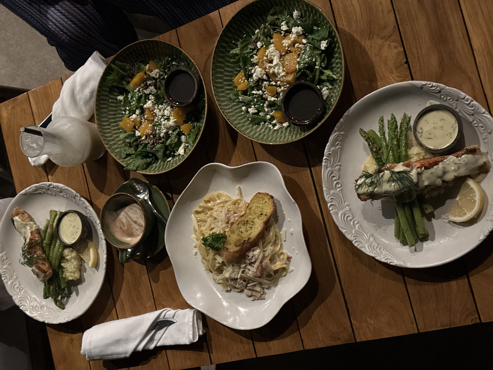 The image shows a wooden table in what appears to be a restaurant setting. There are several plates of food including two salads, a pasta dish, and two servings of fish and asparagus. There is also a pitcher with a light-colored liquid and a cup of coffee. A neatly folded napkin and silverware are present, indicating a place setting. The lighting suggests an indoor dining area, possibly in Nusa Penida, Indonesia, based on the context of the query. No people are visible in the immediate frame. The focus is on the presented food.